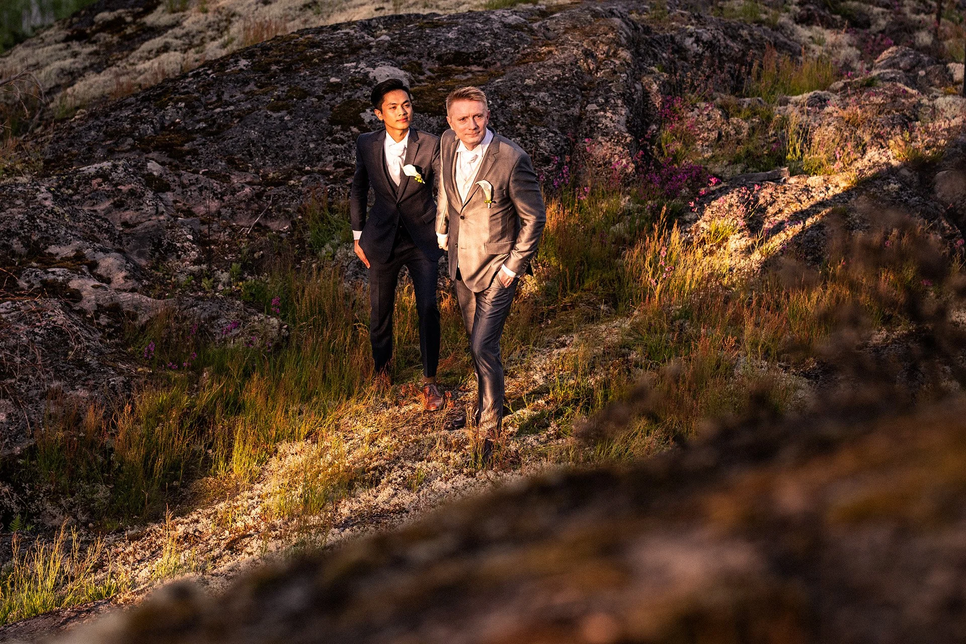 Kaksi miestä juhlapuvuissa kävelee luonnossa iltavalossa, taustalla kallioita ja kasveja.
