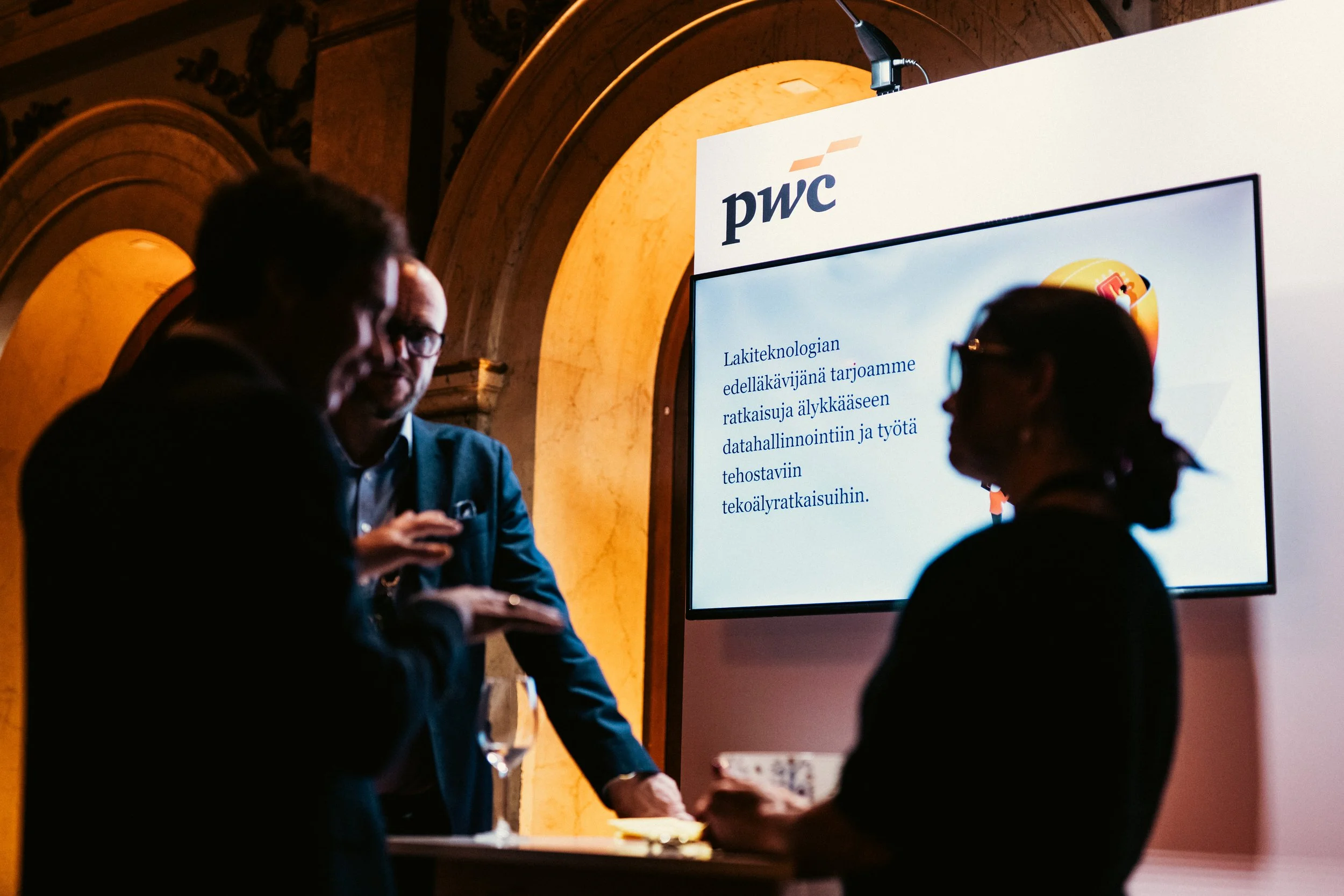 Henkilöitä keskustelemassa konferenssissa, taustalla suuri näyttöruutu, jossa lukee 'pwc' ja suomenkielistä tekstiä.
