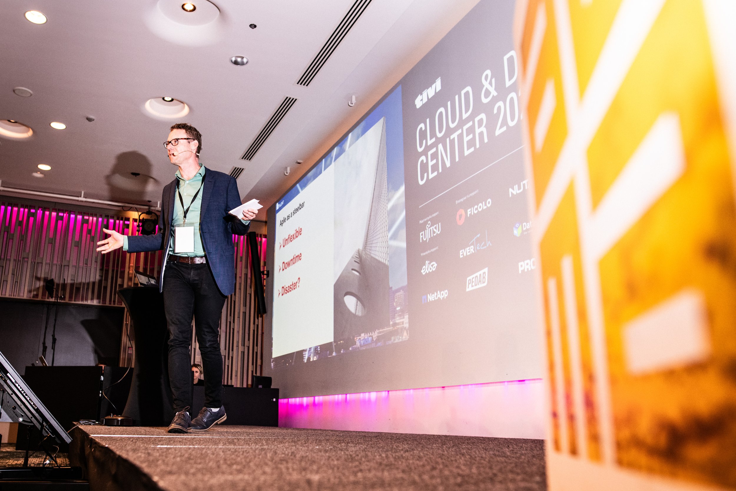 Mies puhuu lavalla esityksen aikana, vieressä iso näyttö, jossa lukee 'CLOUD & D CENTER 20', ja taustalla on värikäs seinä.