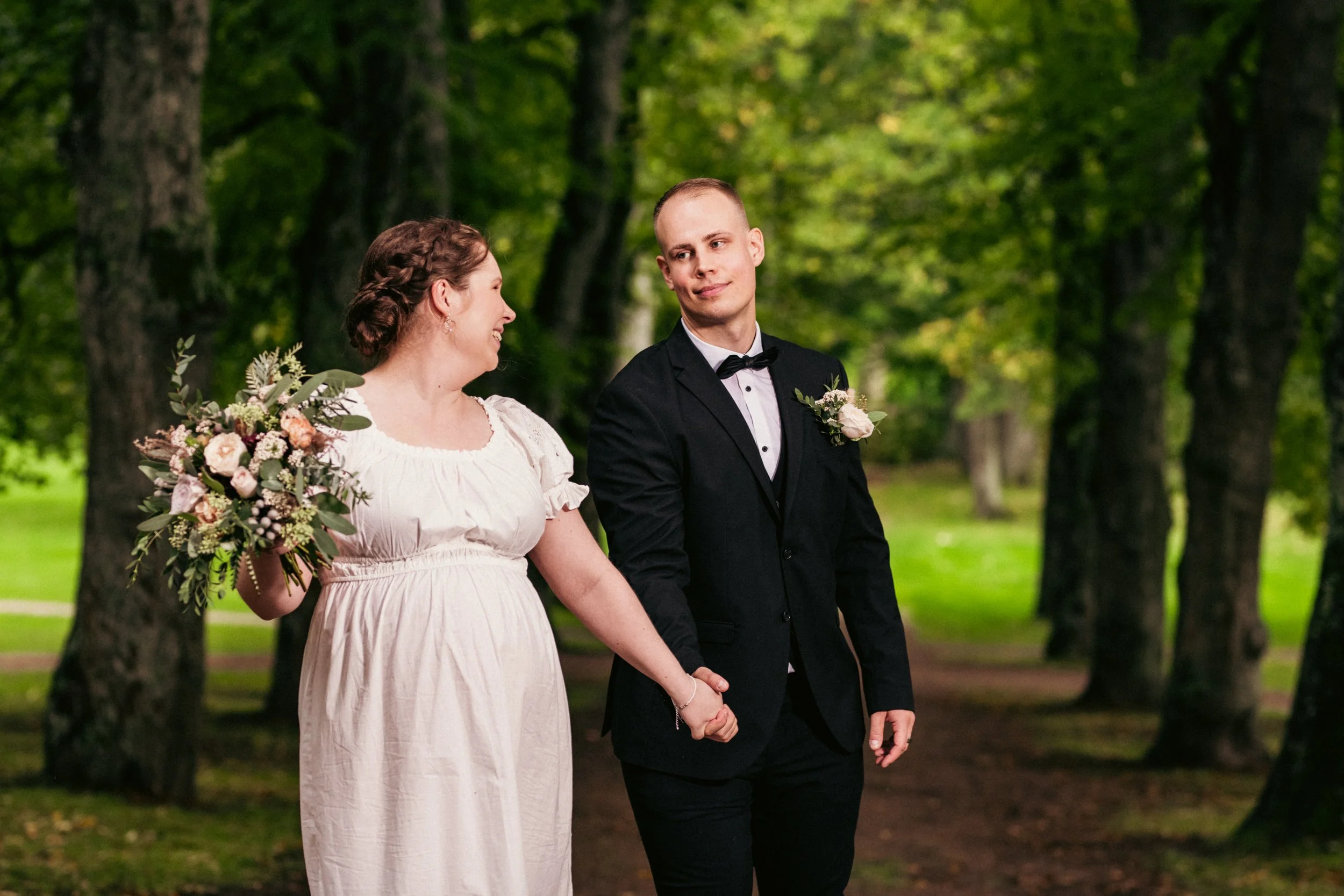 Hääpari kävelee käsipuolessa puistossa, nainen pitää kukkasiltaa ja mies puku ja solmuke.