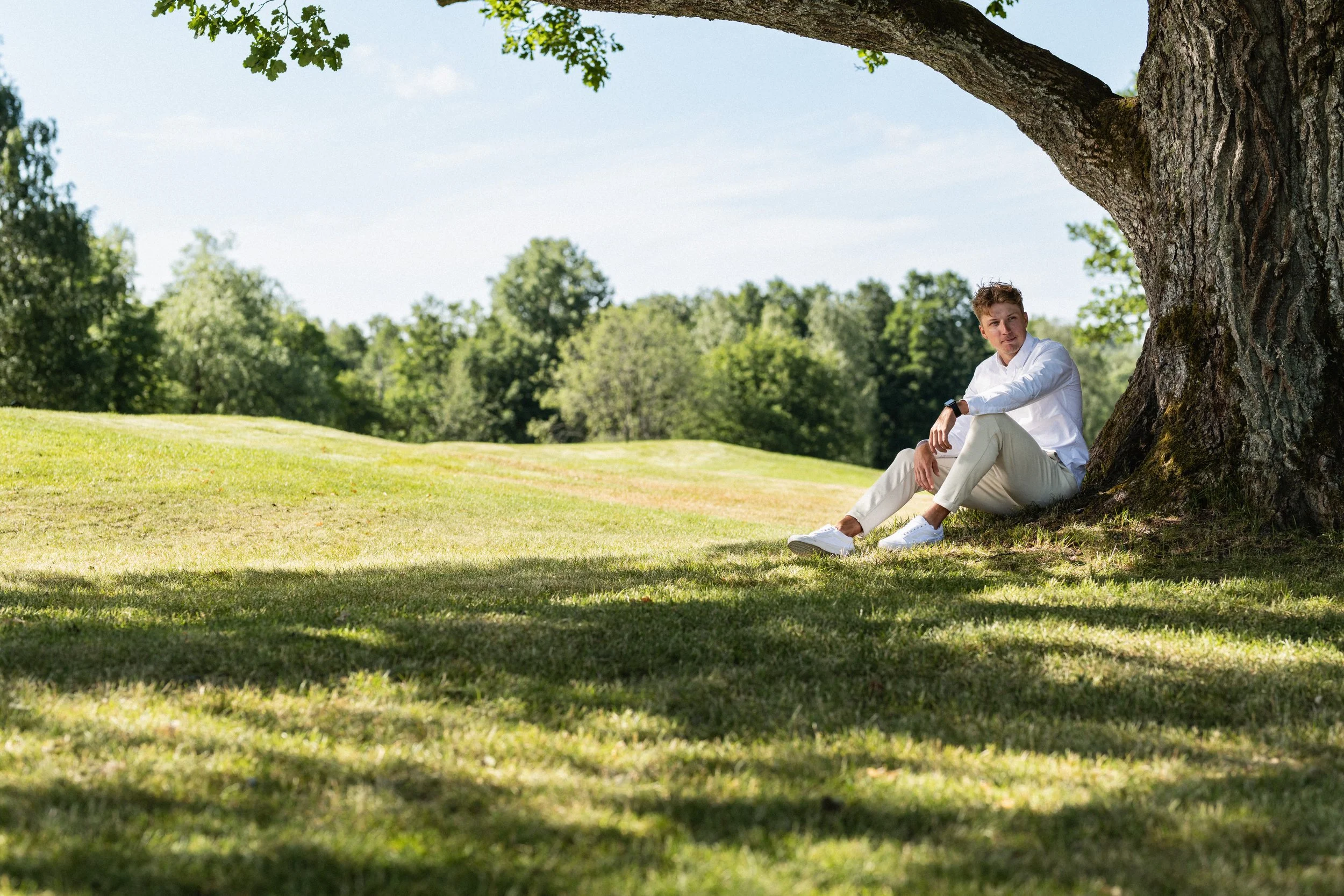 Nuori mies istuu suuren puun varjossa, nurmikkoreunaisella alueella, taustalla puita ja sininen taivas.