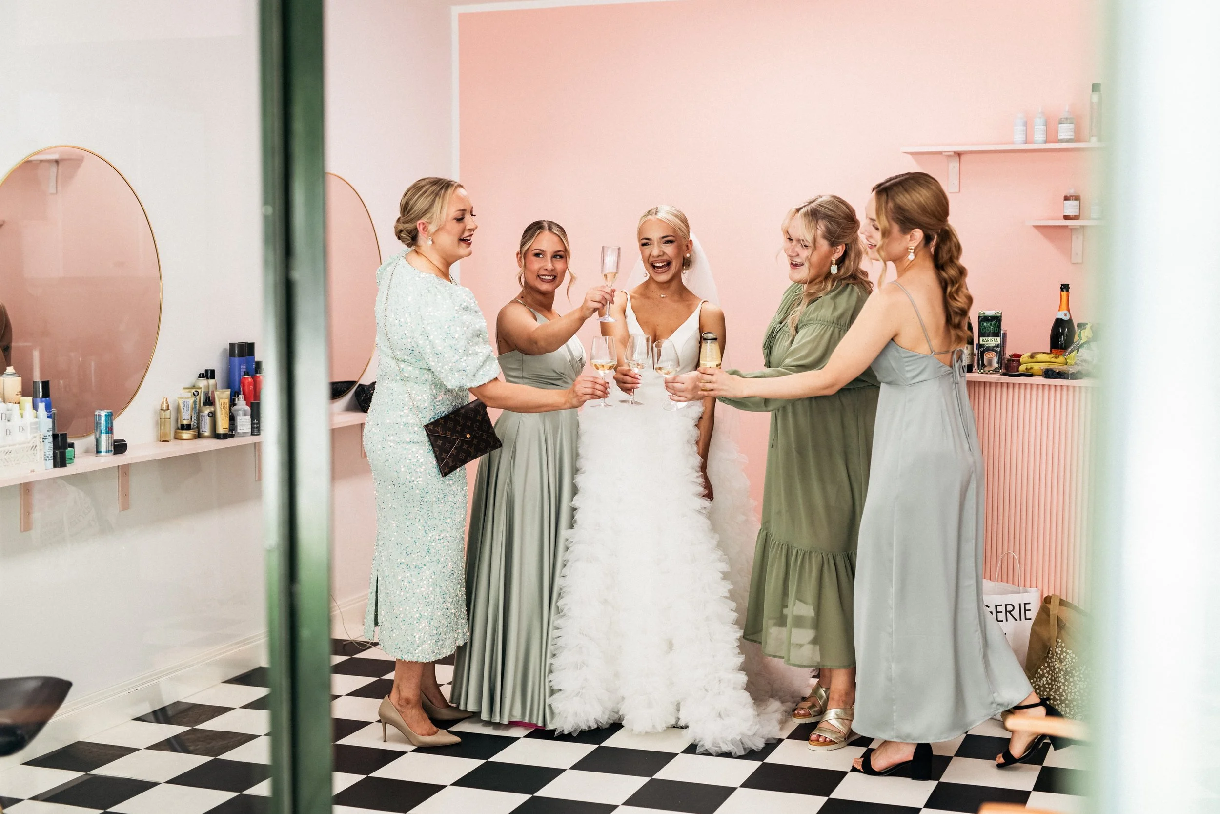 Seitsemän naista juhlivat hääpäivää. Osallistujat kiroilevat maljoja ja nauravat. Taustalla on kosmetiikkapöytä ja peilejä pinkissä huoneessa.