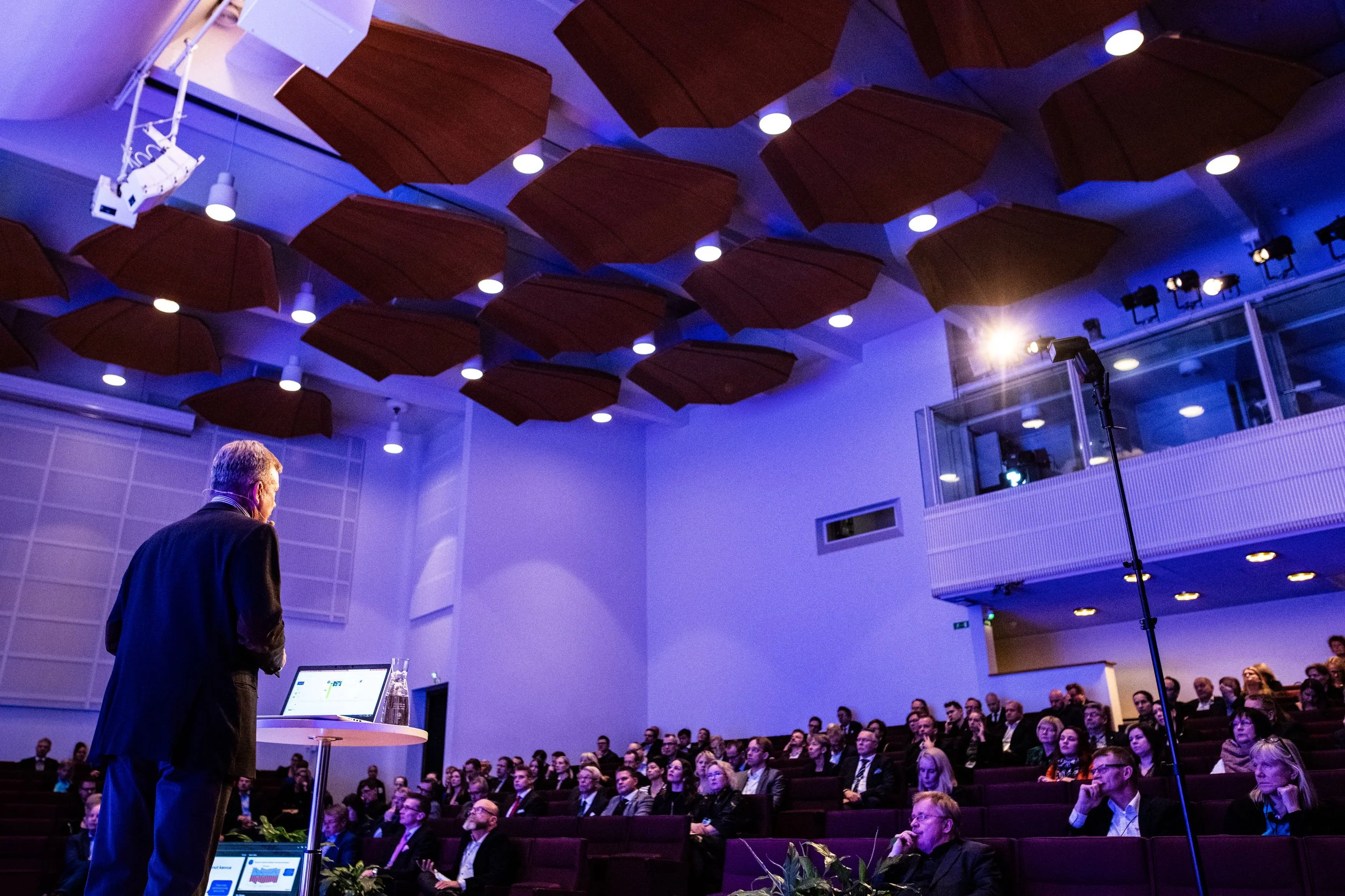 Mies puhuu konferenssipuhetta yleisölle, istuntosalissa, taustalla on katto, jossa on useita puupalikoita ja valoja.