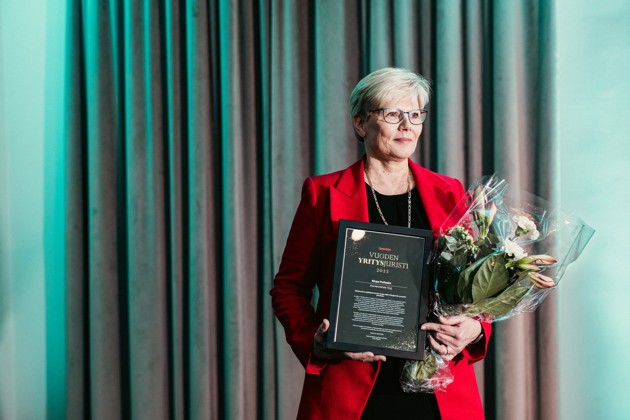 Nainen pikkutakki ja korvakorut, joka pitää palkintolahjakorttia ja kukkavihkoa, seisoo edessä taustalla harmaat verhot.