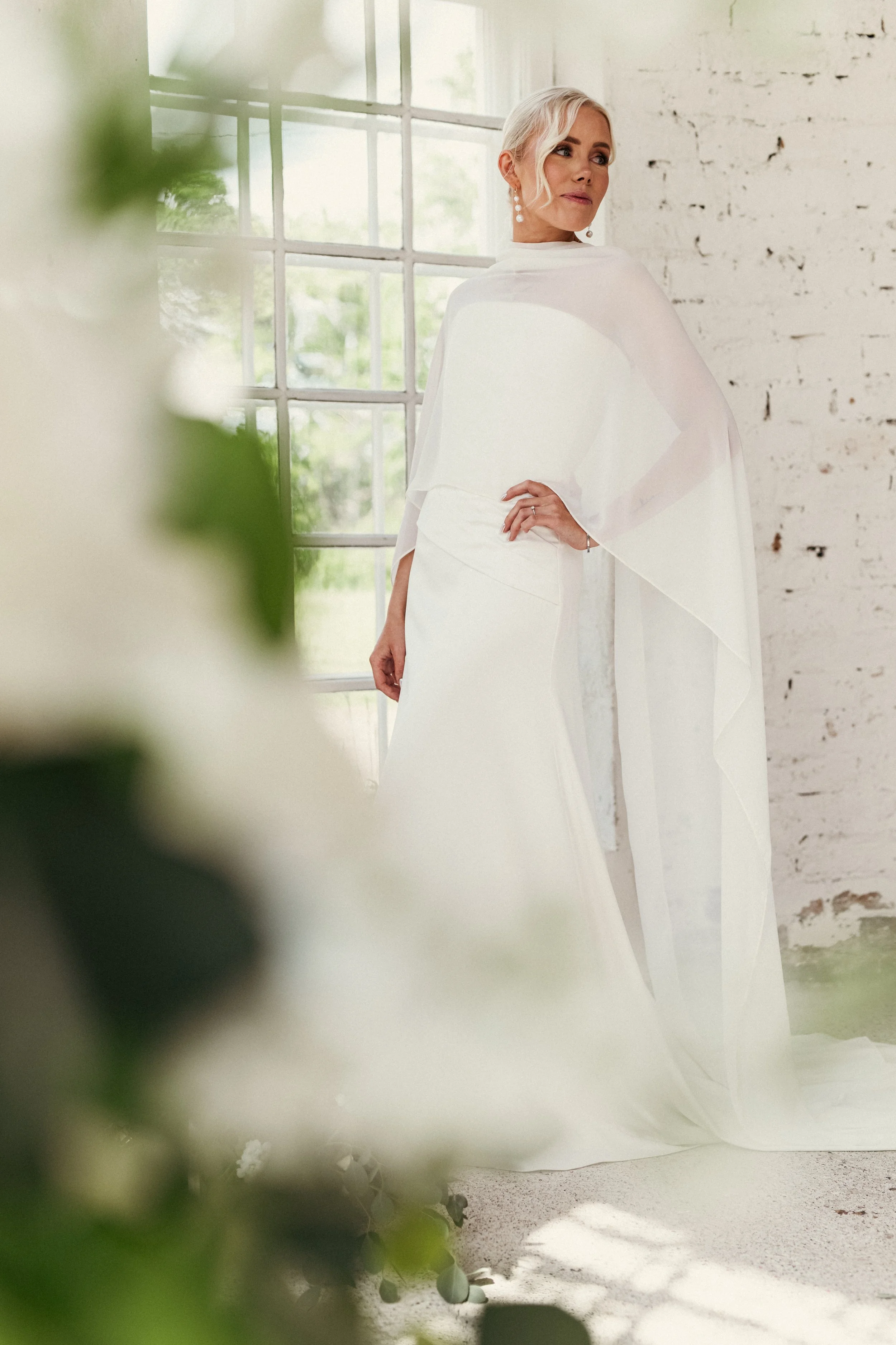 Vanhapioltinen nainen häämekossa seisoo sisällä, taustalla suuri ikkunA ja tiiliseinä.