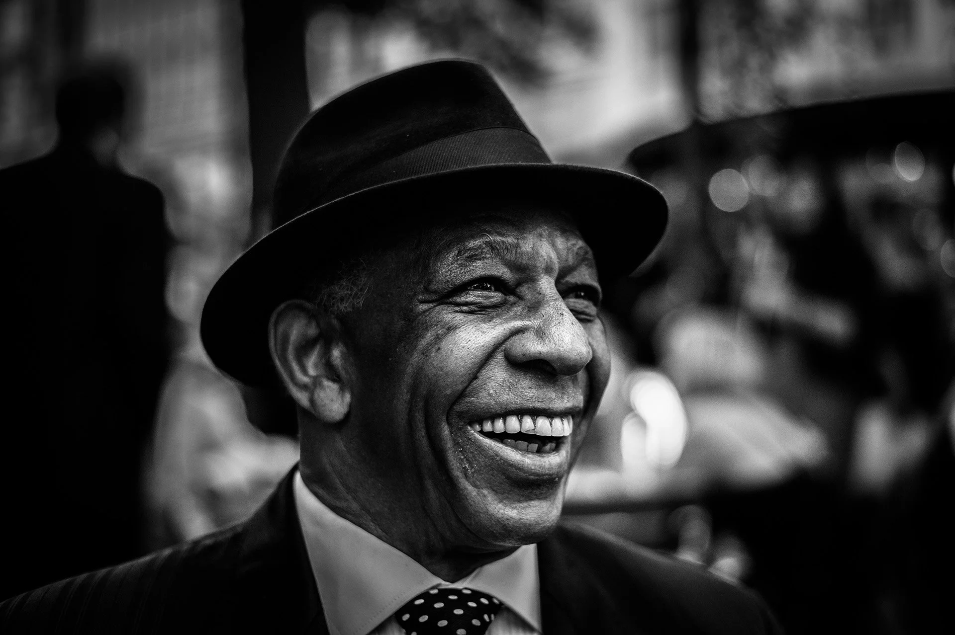 Hymyilevä mies, joka on pukeutunut pukuun ja solmioon, hänellä on hattu päässä, taustalla häilyviä ihmisiä, mustavalkoinen kuva.