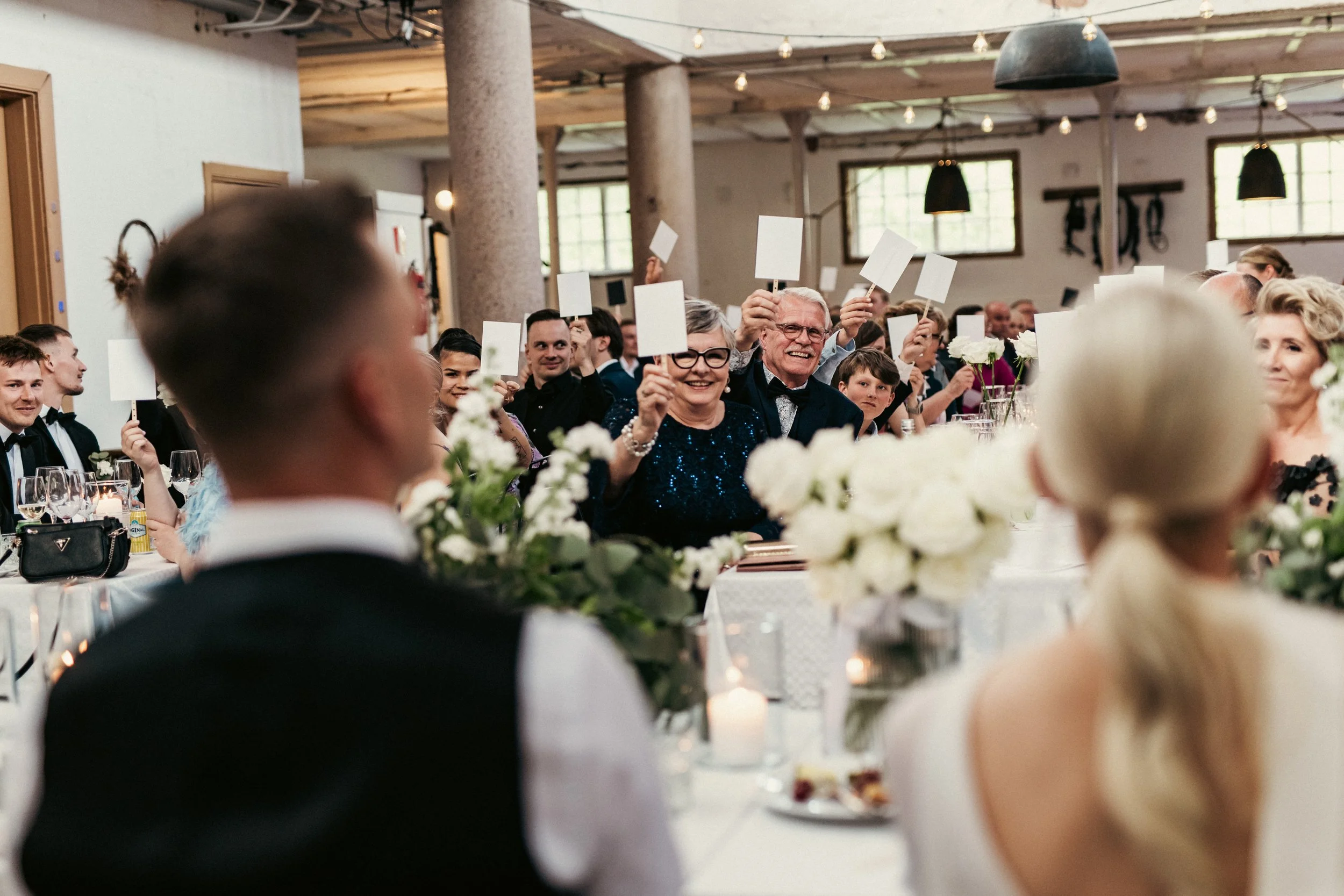 Juhlava ruokapöytä täynnä kukka-asetelmia ja kynttilöitä, täynnä onnellisia ihmisiä, jotka osallistuvat kuin leikkimieliseen peliin, jossa he pitävät pieniä valkoisia lippuja. Taustalla vanhanaikaisia ikkunoita ja valaisimia, ja tunnistamaton pari hu