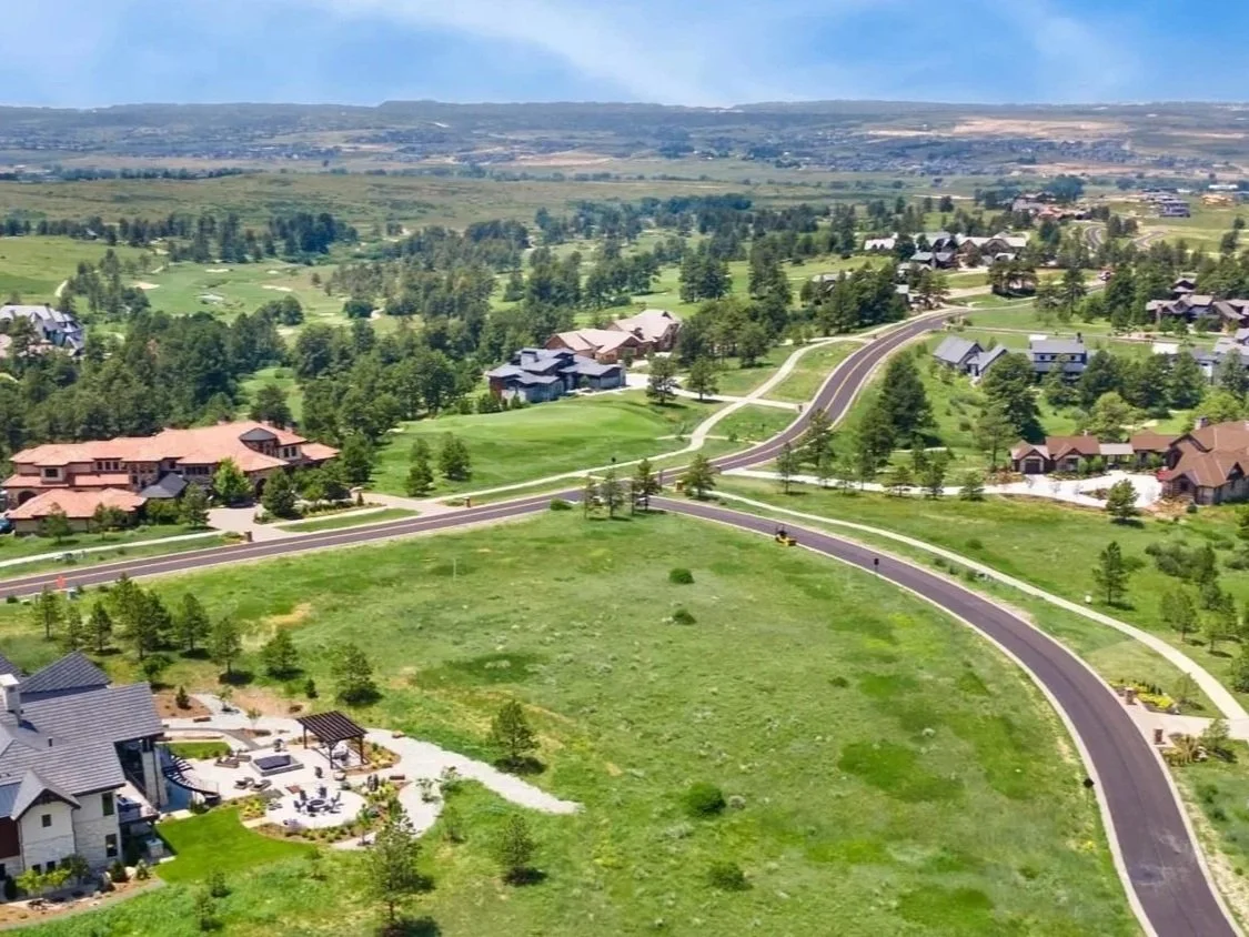 Aerial view of a suburban neighborhood with houses, roads, green lawns, and trees surrounded by open fields and distant hills under a partly cloudy sky.