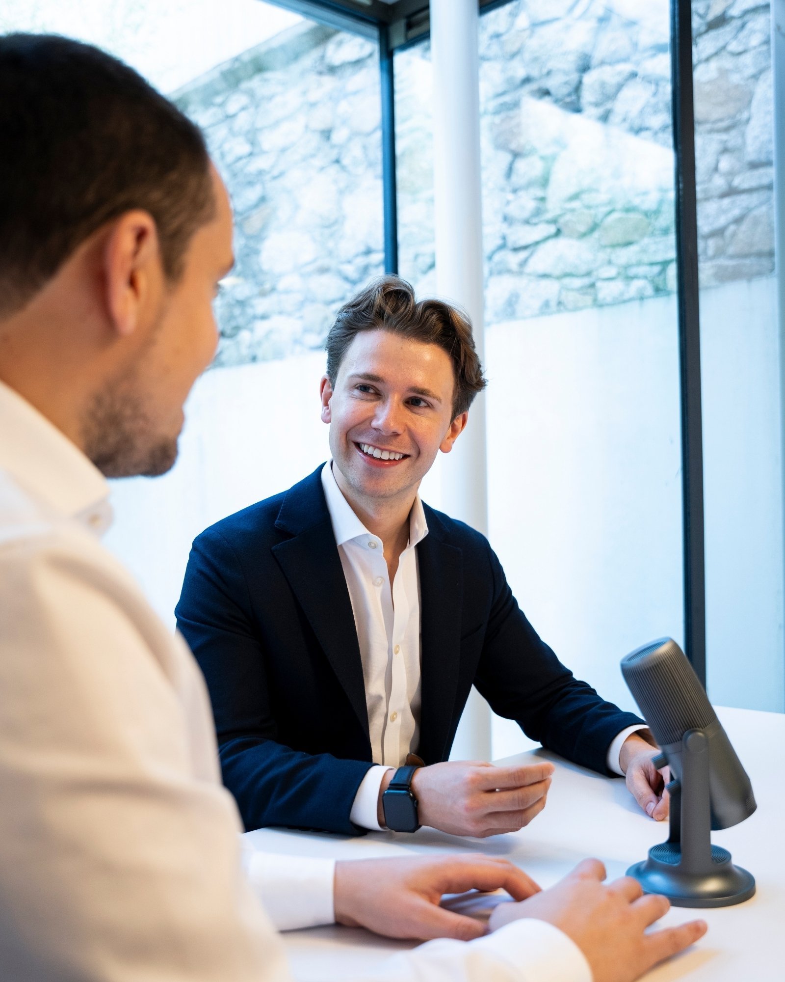 Dale McDermott sitting at table with a microphone recording podcast, inside a glass-walled room with a stone wall visible outside.