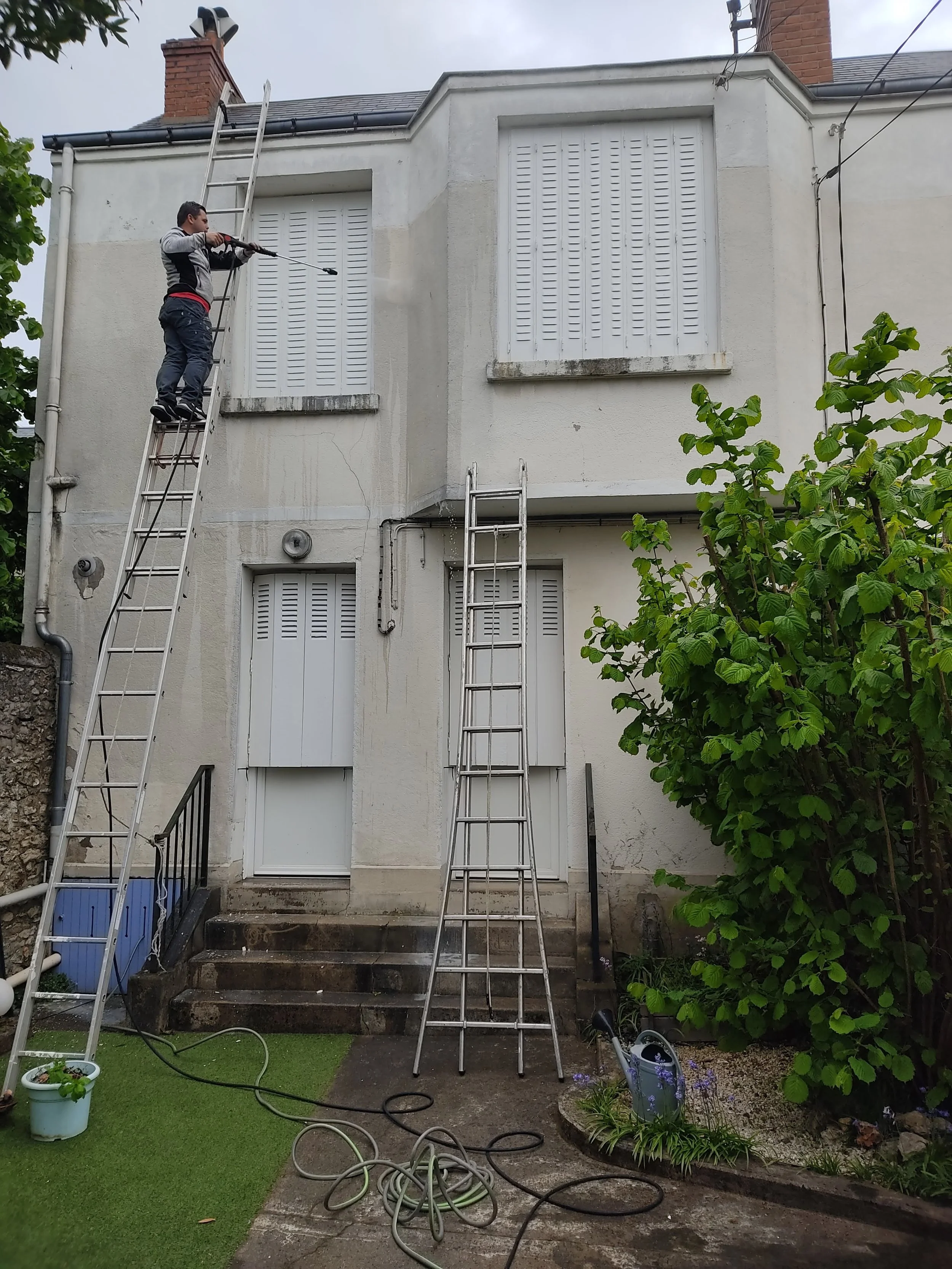 Une personne utilisant une perceuse pour réparer ou nettoyer une fenêtre à l'étage d'une maison, avec une échelle de taille moyenne et des volets fermés, dans un jardin avec des plantes et un tuyau d'arrosage au sol.