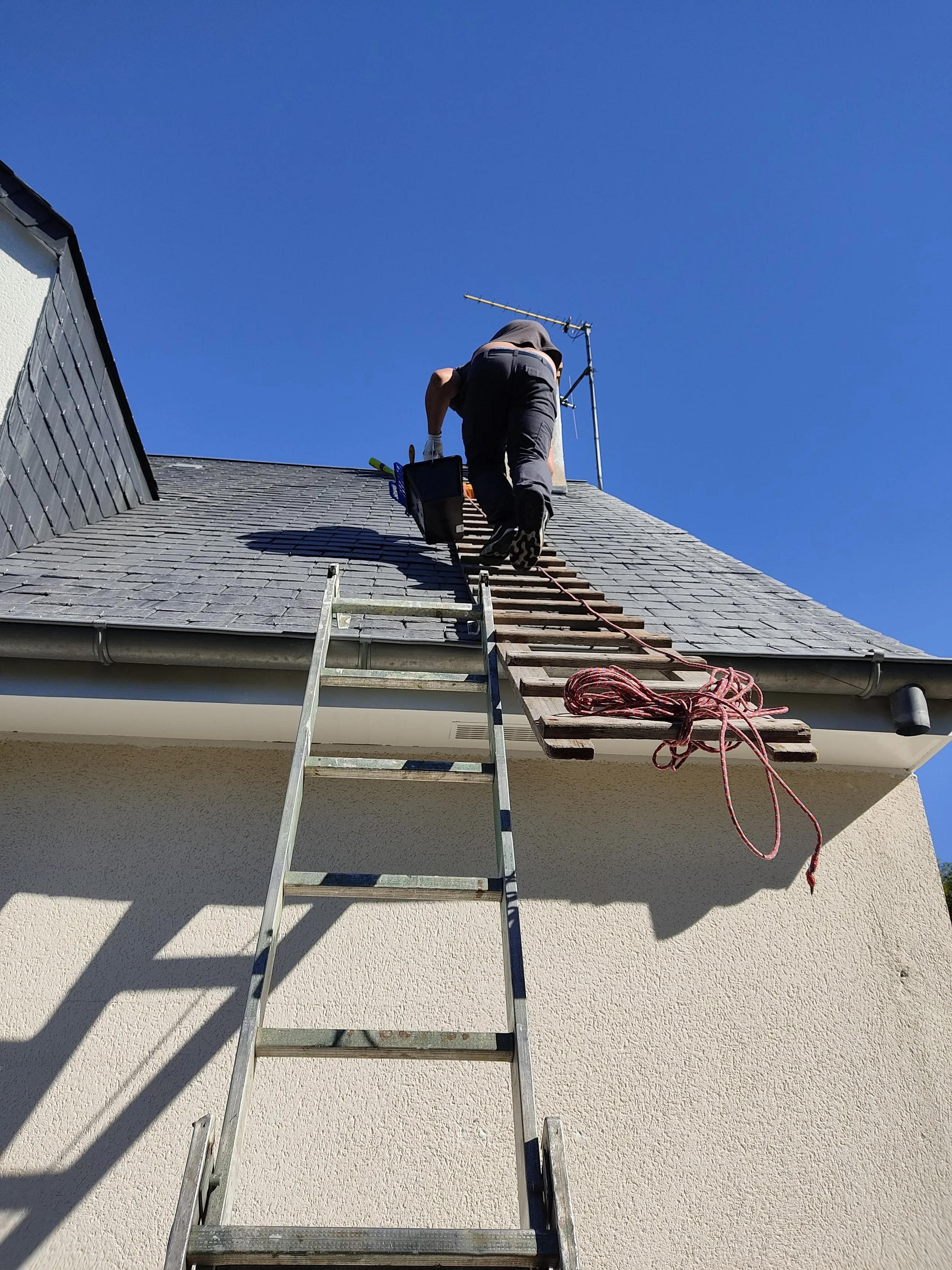 Un homme grimpe une échelle pour accéder au toit d'une maison, avec des outils et une corde à proximité, sous un ciel bleu clair.