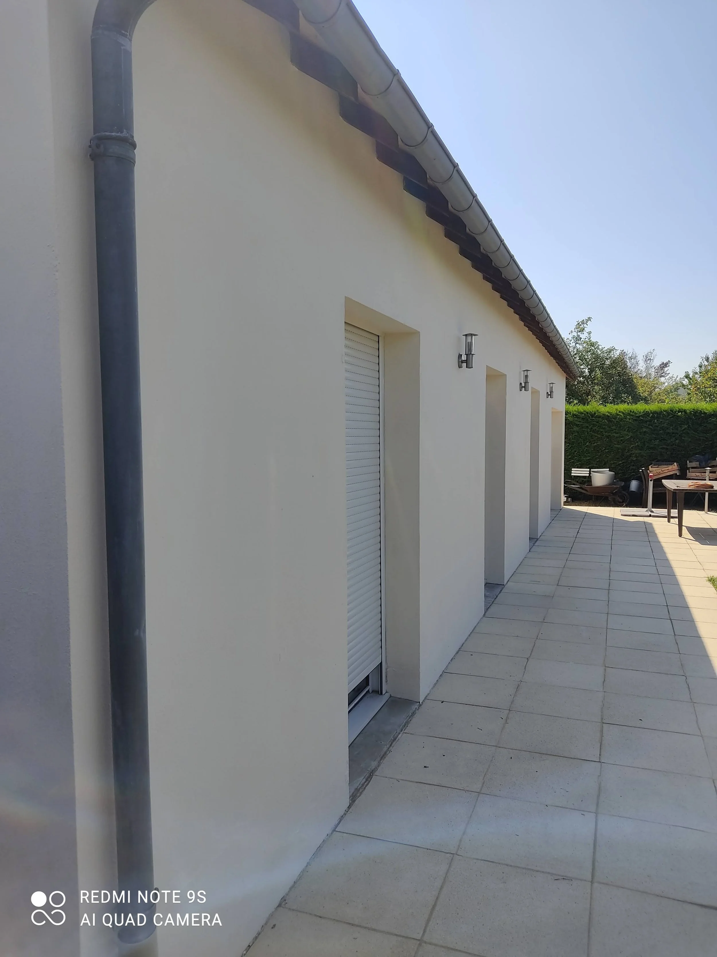 Façade d'une maison moderne avec un mur blanc, des volets roulants, un éclairage extérieur et une terrasse pavée, dans un jardin ensoleillé.