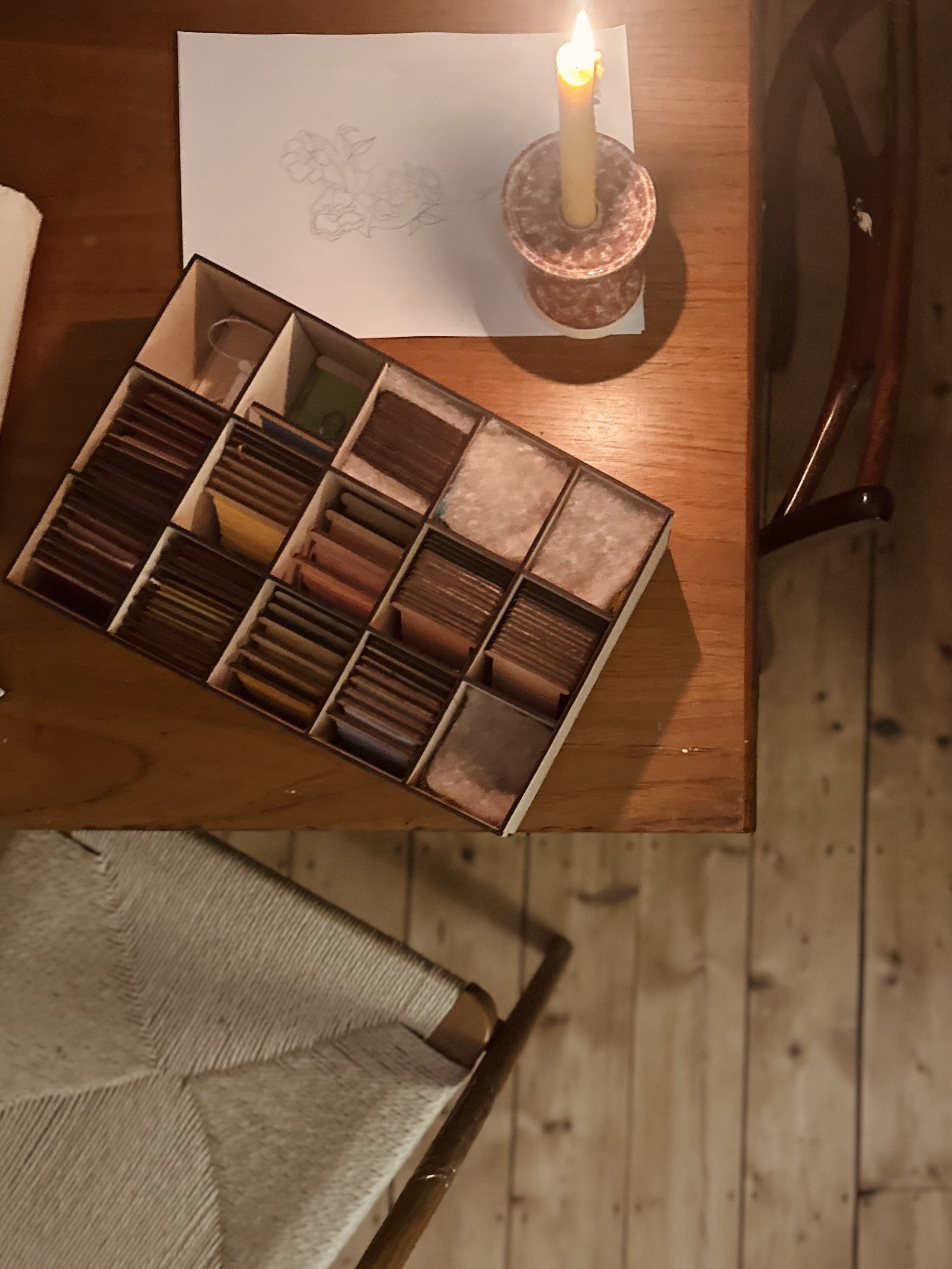 The image is a birds eye view shows a wooden box filled with wooden embroidery cards, the coloured threads just visible. The box is on top of a rectangular teak table and is lit by a beeswax candle sitting in a ceramic candle holder.