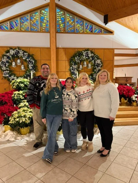 Family of five posing in front of Christmas wreaths and poinsettias in a church or decorated hall.
