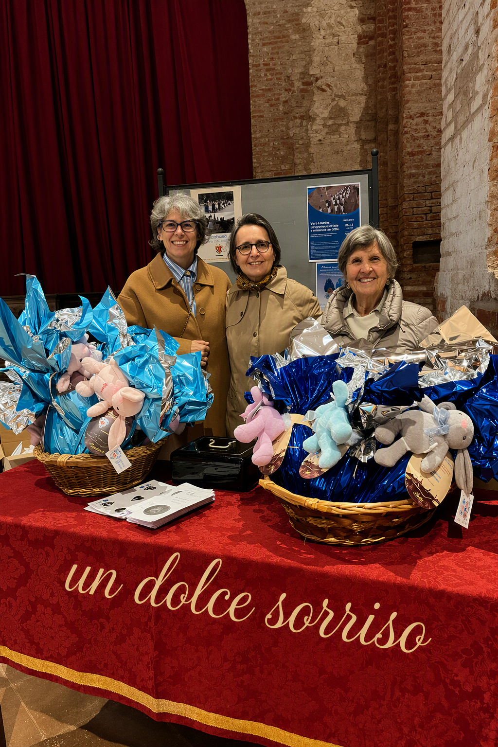 Tre donne sorridenti dietro a un tavolo con regali di peluche, con un tavolo coperto da una tovaglia rossa, la scritta 'un dolce sorriso'. Gli scaffali di mattoni visibili sullo sfondo.
