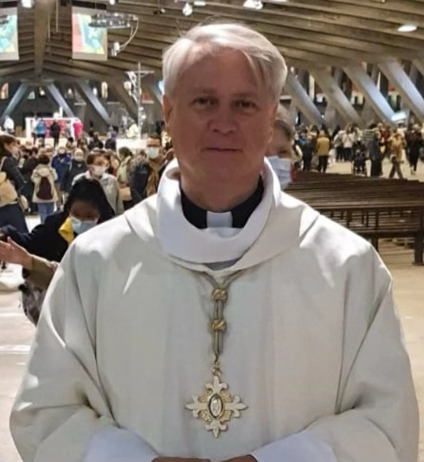 Un sacerdote vestito con abiti religiosi bianco e un ciondolo con croce d'oro e immagine al centro. Sullo sfondo, una chiesa o un luogo di culto con molte persone presenti, alcune con mascherina.