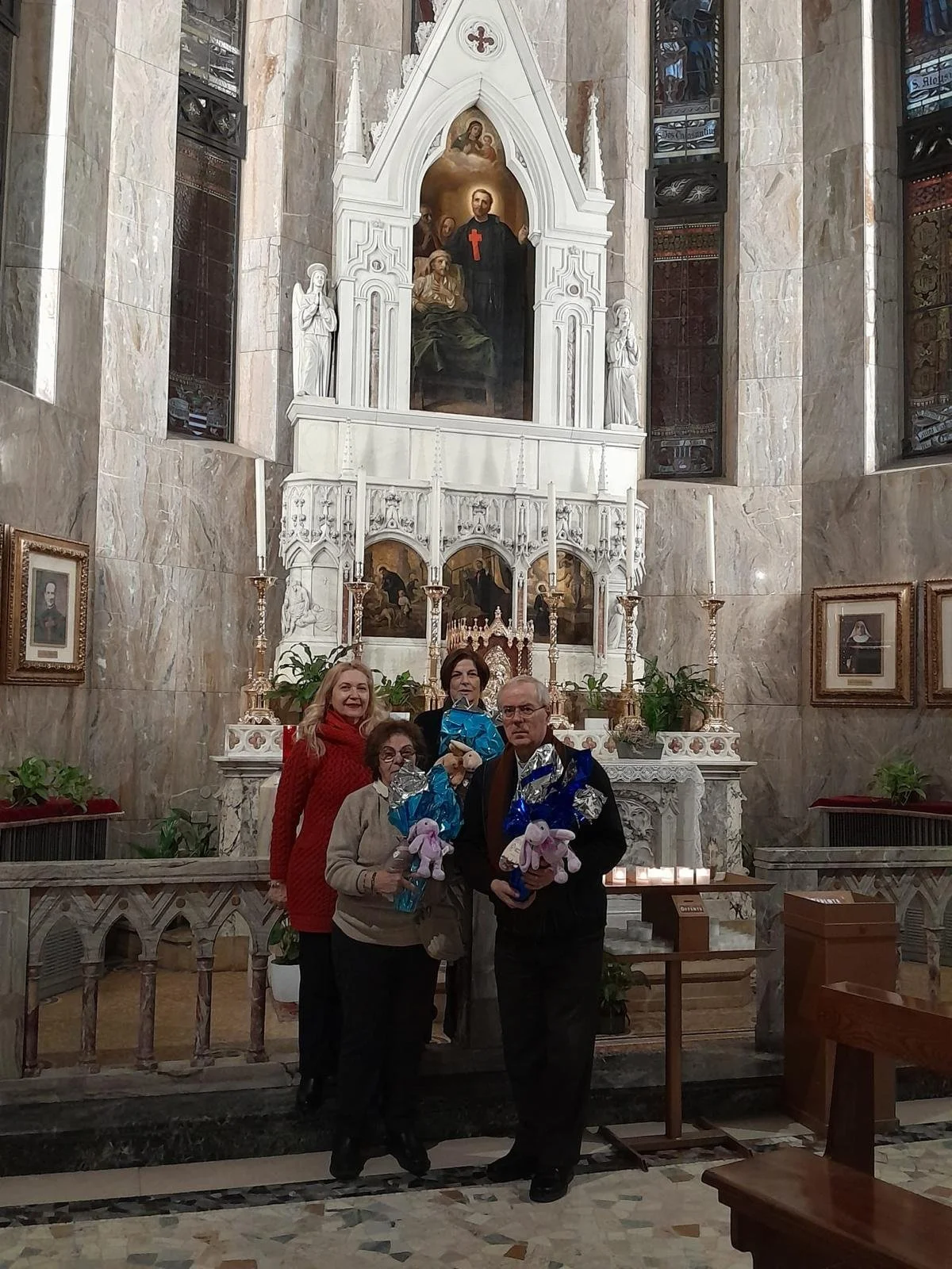Quattro persone, due donne anziane e due più giovani, all'interno di una chiesa antica, che tengono dei peluche e si trovano davanti a un altare decorato con candele e piante, con un grande dipinto religioso sullo sfondo.