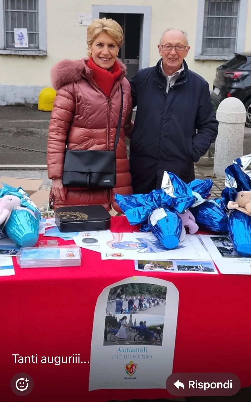 Due persone sorridenti davanti a un banco con doni e volantini, con scritte in italiano, in un'area esterna.