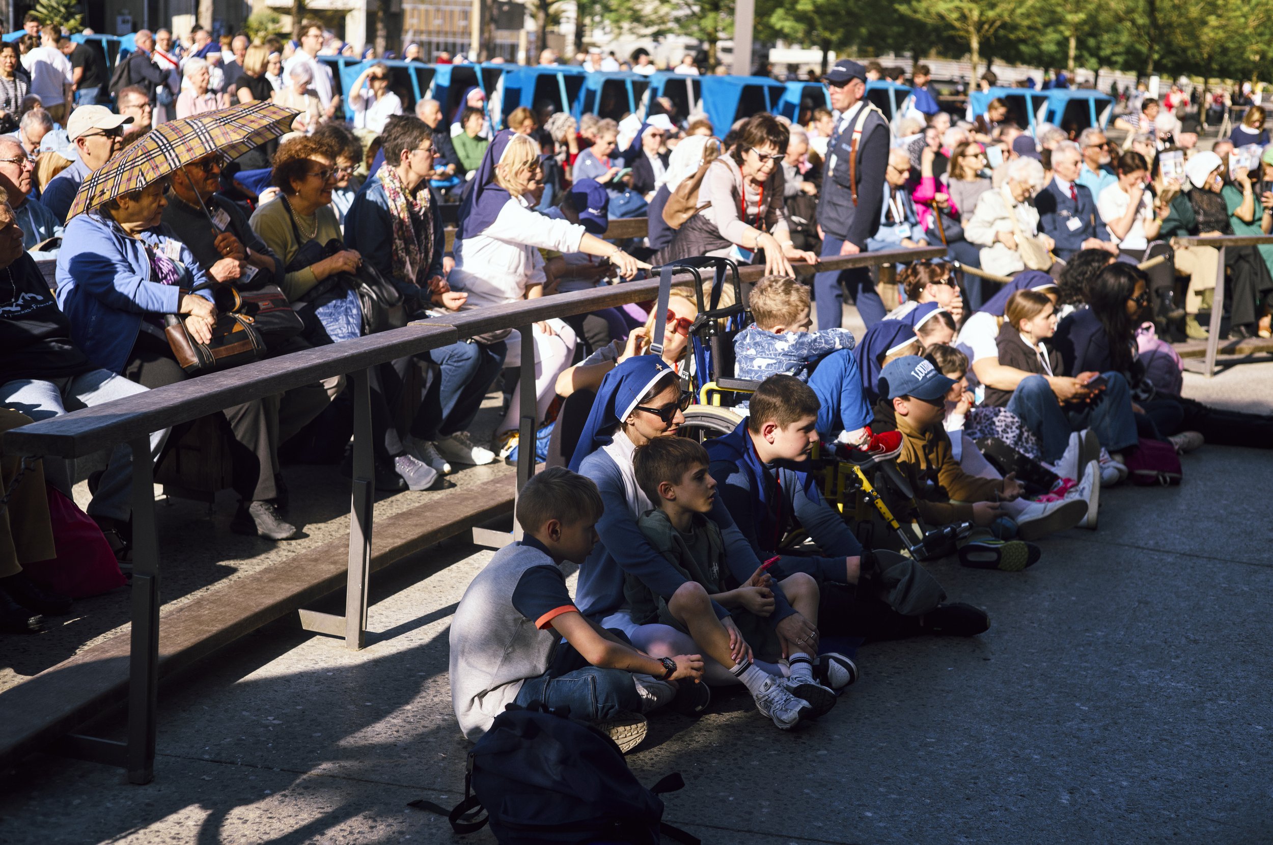 Gruppo di persone sedute su una recinzione di legno in attesa di un evento, con alcune persone che si riparano dal sole con un ombrello. In sfondo ci sono autobus blu e alberi.