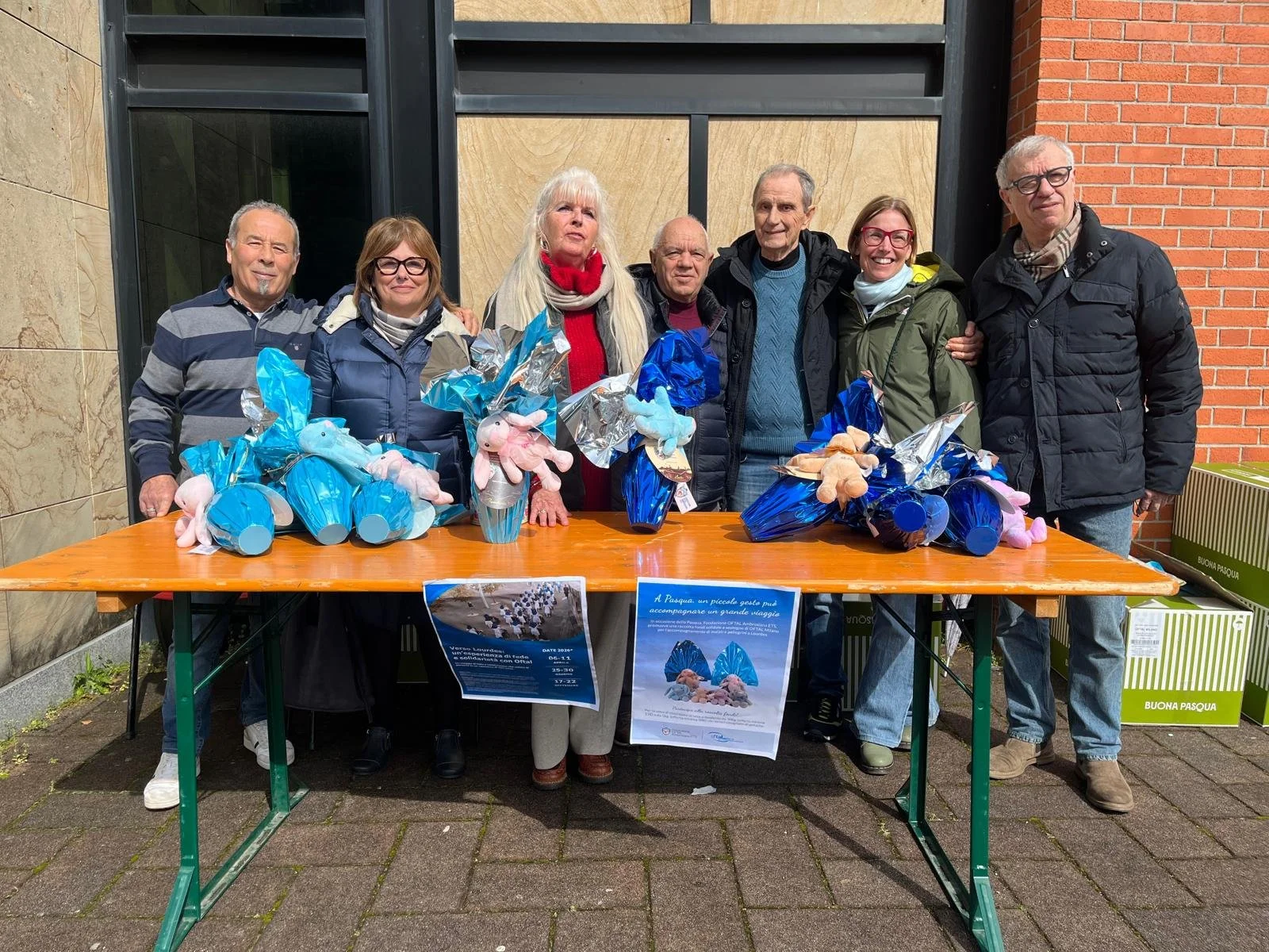 Gruppo di persone che donano dei pulcini di peluche e confezioni decorate con plastica blu, in occasione della Pasqua, davanti a una tavola con due poster informativi.