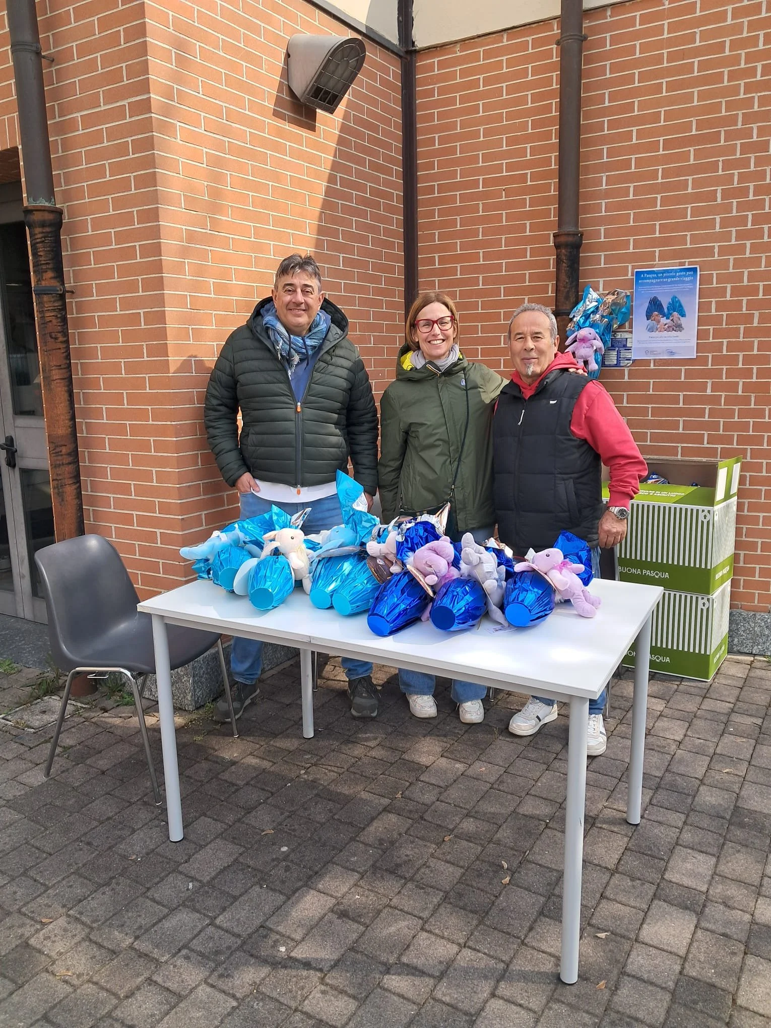 Tre persone sorridenti davanti a un tavolo con conigli di peluche in schiuma blu e rosa, con sacchetti di pasqua e un cartellone che ricorda la Pasqua, all'aperto davanti a un edificio con mura di mattoni.