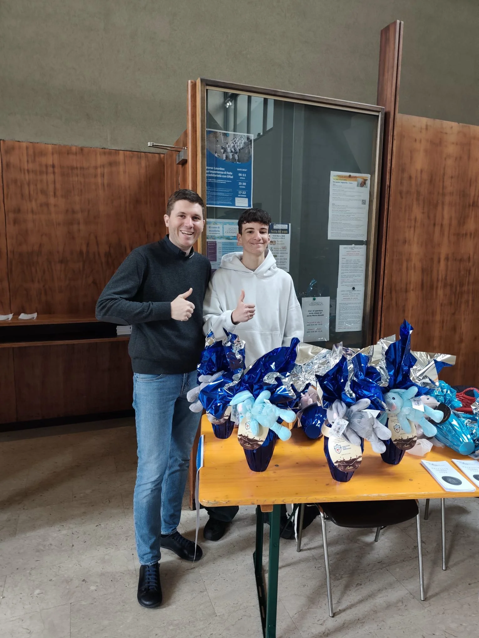 Due uomini sorridenti davanti a un tavolo con decorazioni blu e argento, in un ambiente interno con pannelli di legno e poster alle pareti