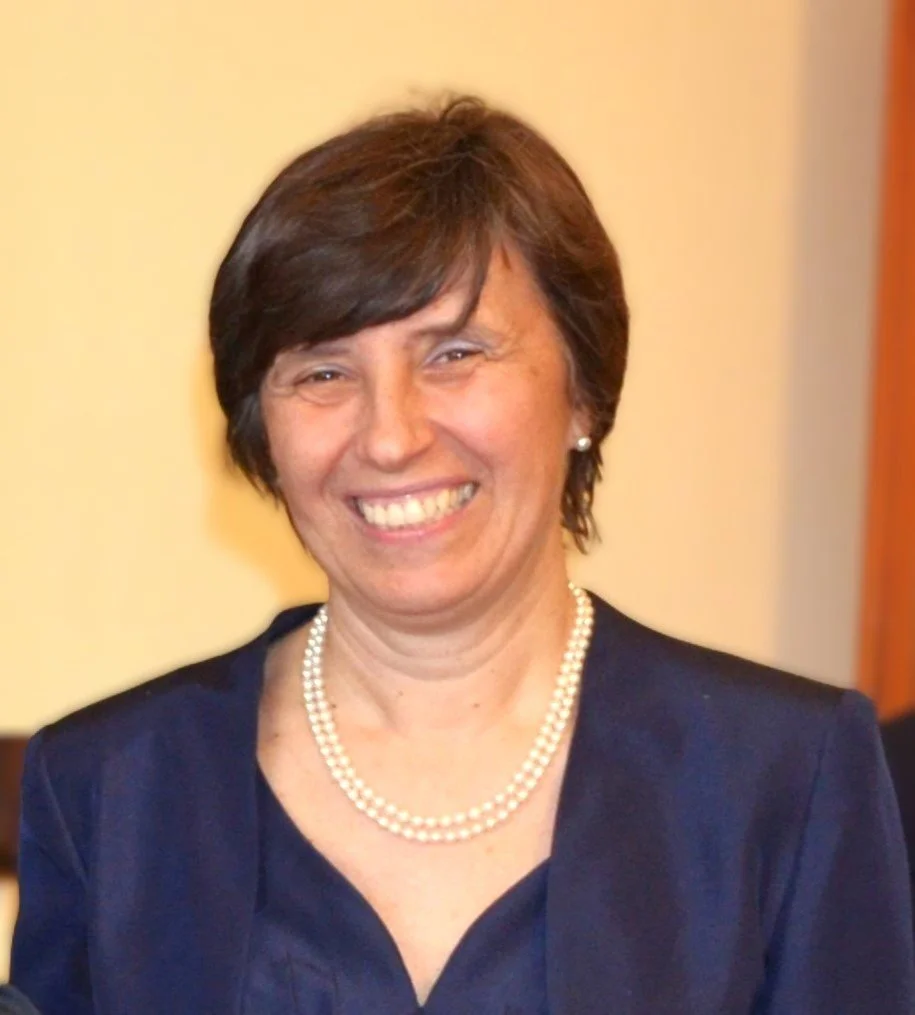 Donna sorridente con abiti eleganti, collana di perle, capelli corti castani, sfondo neutro.