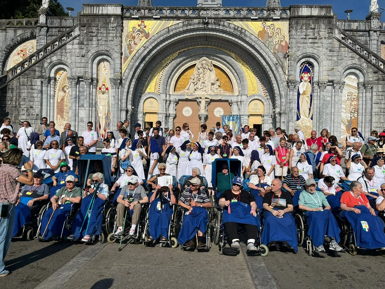 Un gruppo di persone, alcune in sedia a rotelle e altri in piedi, davanti a una chiesa decorata con mosaici e sculture. È una giornata di sole con molti partecipanti a un evento.