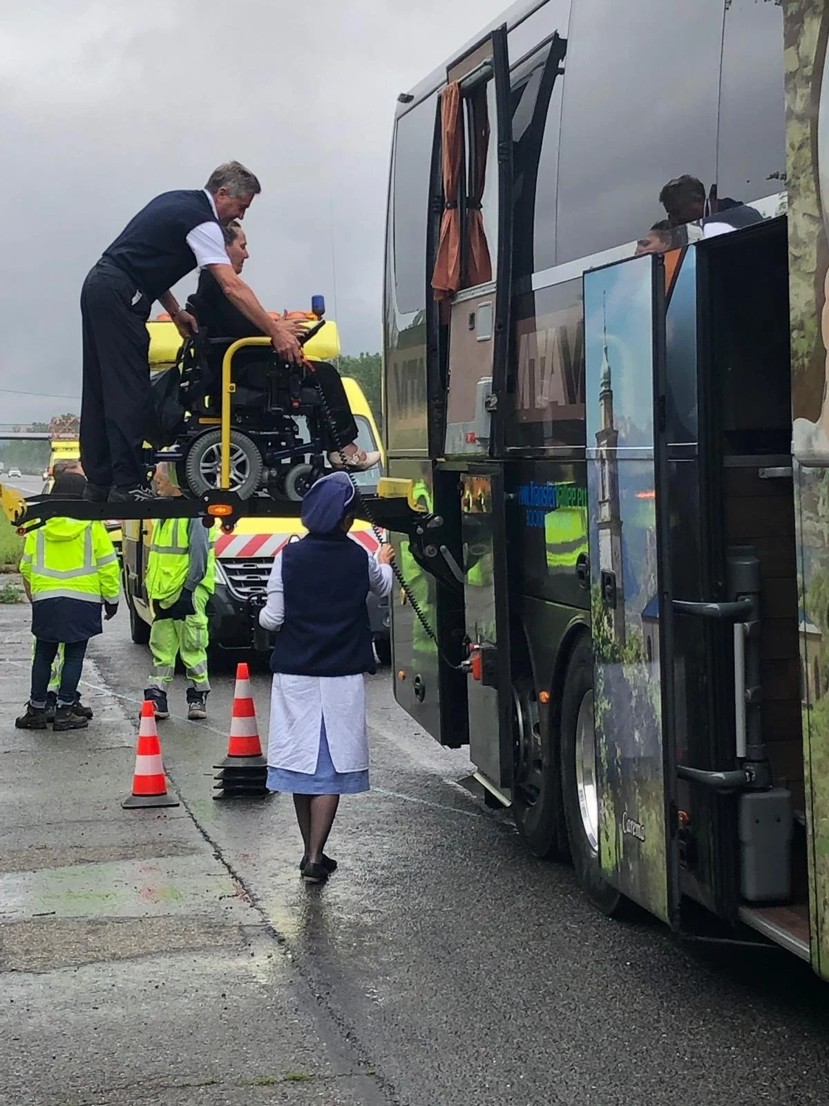 Operatori tra cui una donna in divisa, assistono una persona in sedia a rotelle che viene caricata su un autobus con un sollevatore. Ci sono altri operatori in alta visibilità e con palanche di traffico vicino, in una giornata nuvolosa.