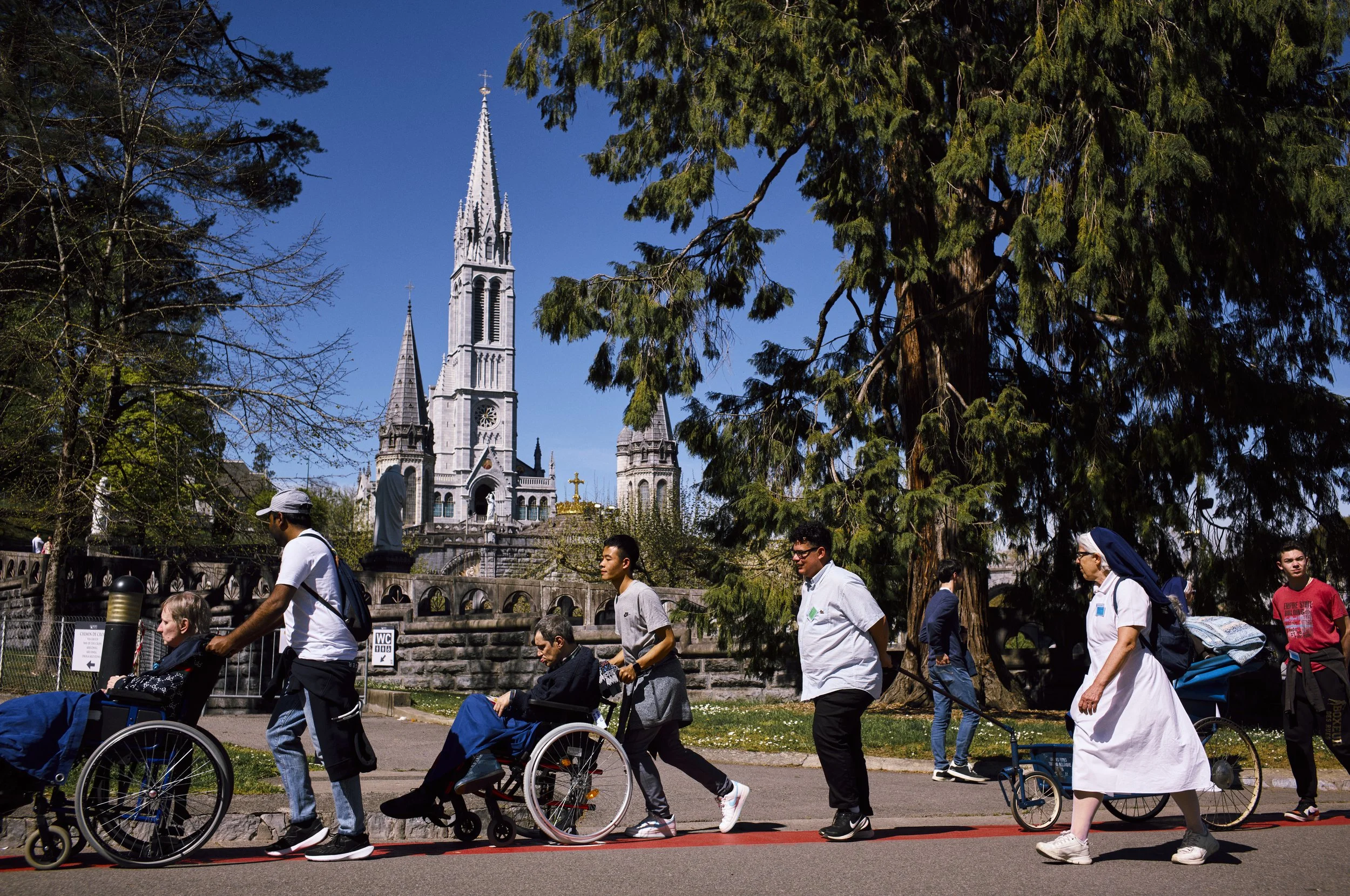 Gruppo di persone con disabilità che passeggiano in un parco, con alcuni in sedia a rotelle e altre che camminano, mentre sullo sfondo si vede un edificio ecclesiastico con alte torri e un cielo sereno.