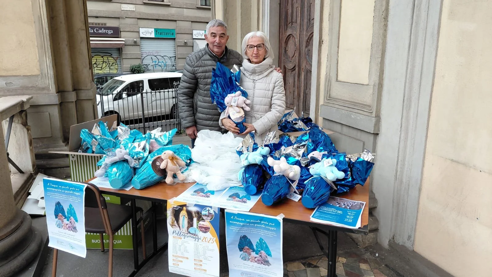 Due persone stanno vendendo coniglietti di cioccolato diPasqua su un tavolo all'aperto, con volantini e decorazioni blu e argento, davanti a un edificio storico.