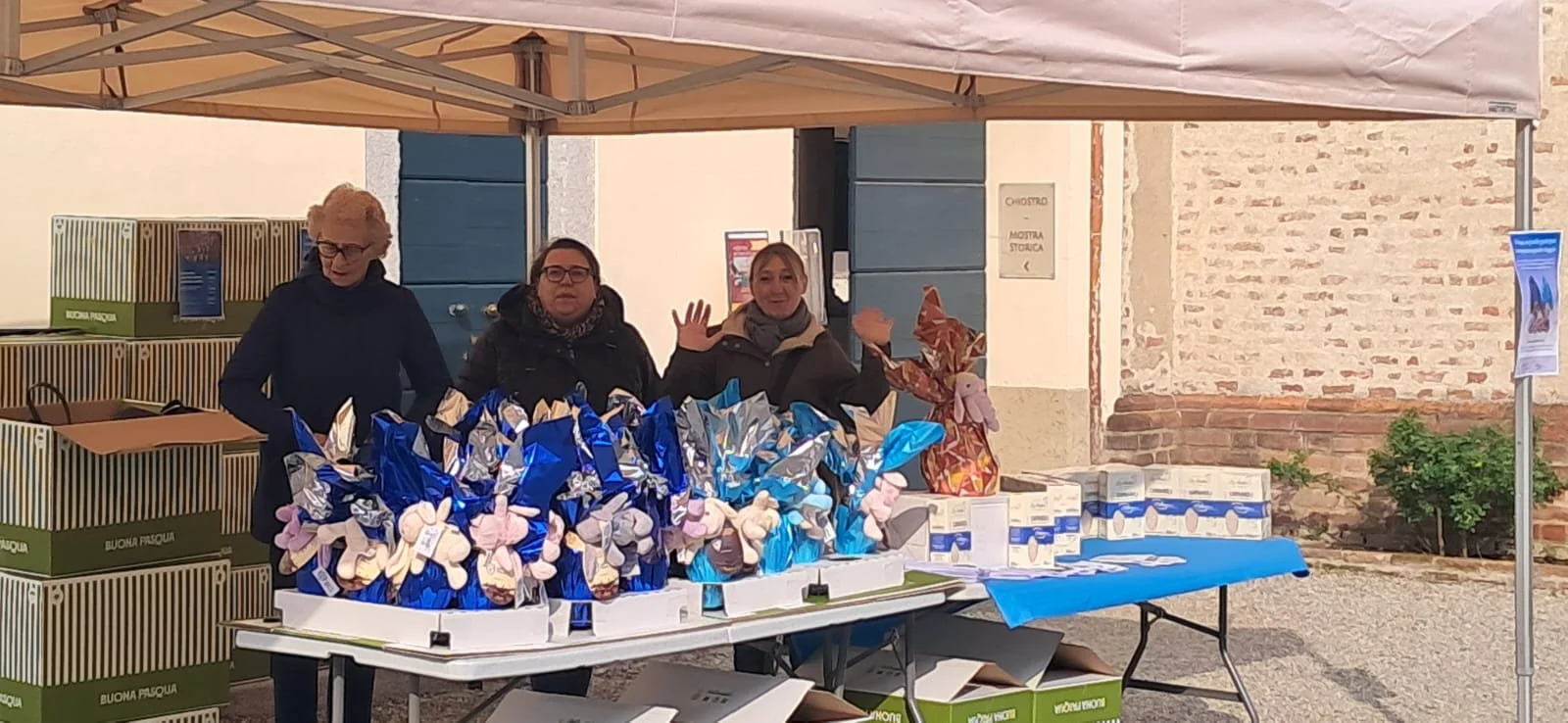 Tre donne dietro un banco di una bancarella con prodotti alimentari, sotto una copertura di colori beige, con scatole verdi e altri prodotti visibili.