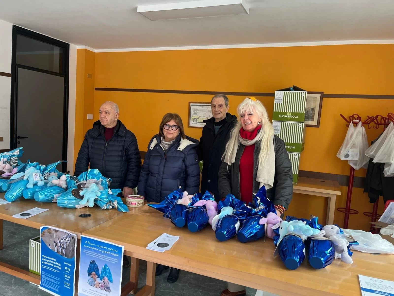 Quattro persone stanno dietro un tavolo con ovetti di Pasqua e conigli di peluche. C'è una parete arancione e vari poster e scatole sullo sfondo.