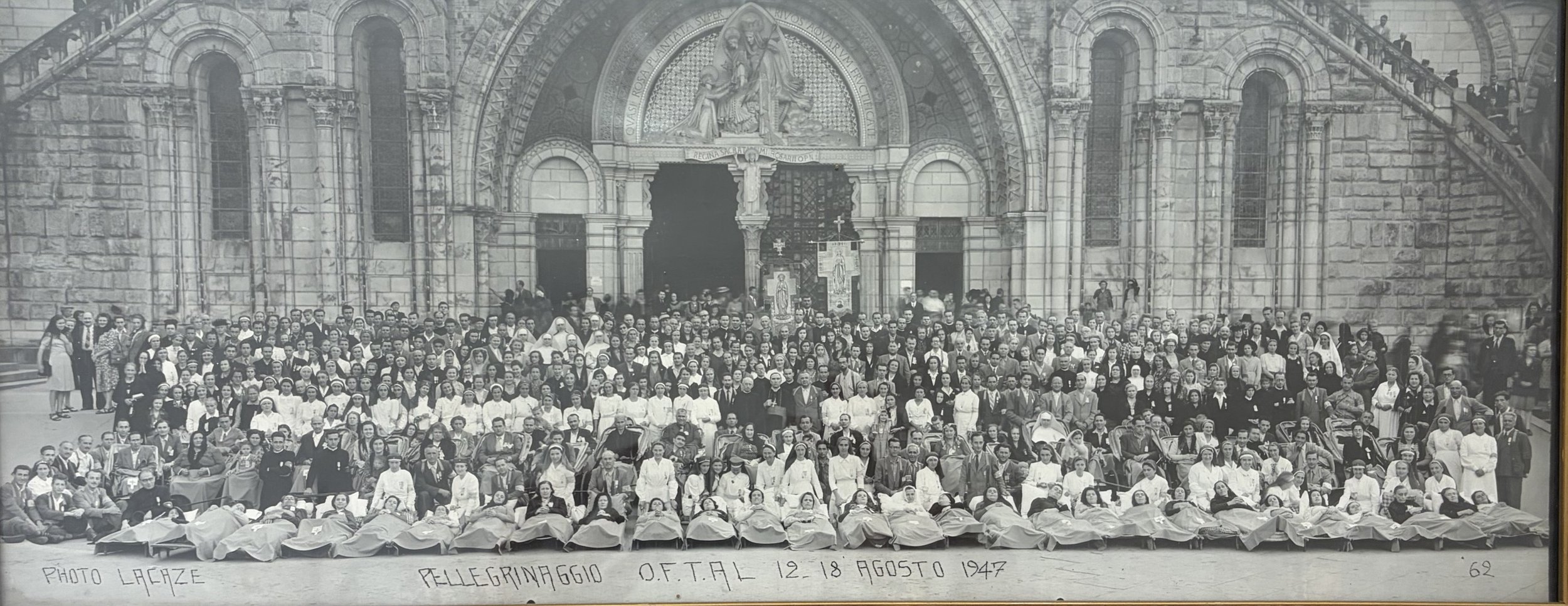 Una grande foto di gruppo davanti a una chiesa, con molte persone in piedi e alcune sdraiate a terra, probabilmente di medici o infermieri, secondo l'abbigliamento. Sullo sfondo, una chiesa con facciata ricca di dettagli architettonici e un'immagine religiosa sopra l'ingresso. Alla base della foto, una scritta in italiano indica che questa foto risale al 18 agosto 1947, con didascalia 'Foto Lañez Pellegrinaggio O.F.T.A.L. 12.18 Agosto 1947'.