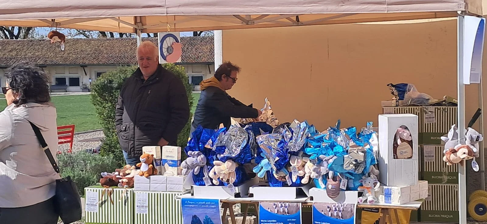 Stanza di mercato con venditori e clienti sotto un tendone, che vende prodotti con decorazioni pasquali