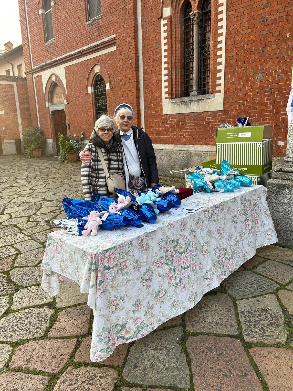 Due donne anziane sorridenti dietro un tavolo con coniglietti di peluche e pacchi regalo blu, davanti a una chiesa di mattoni rossi.