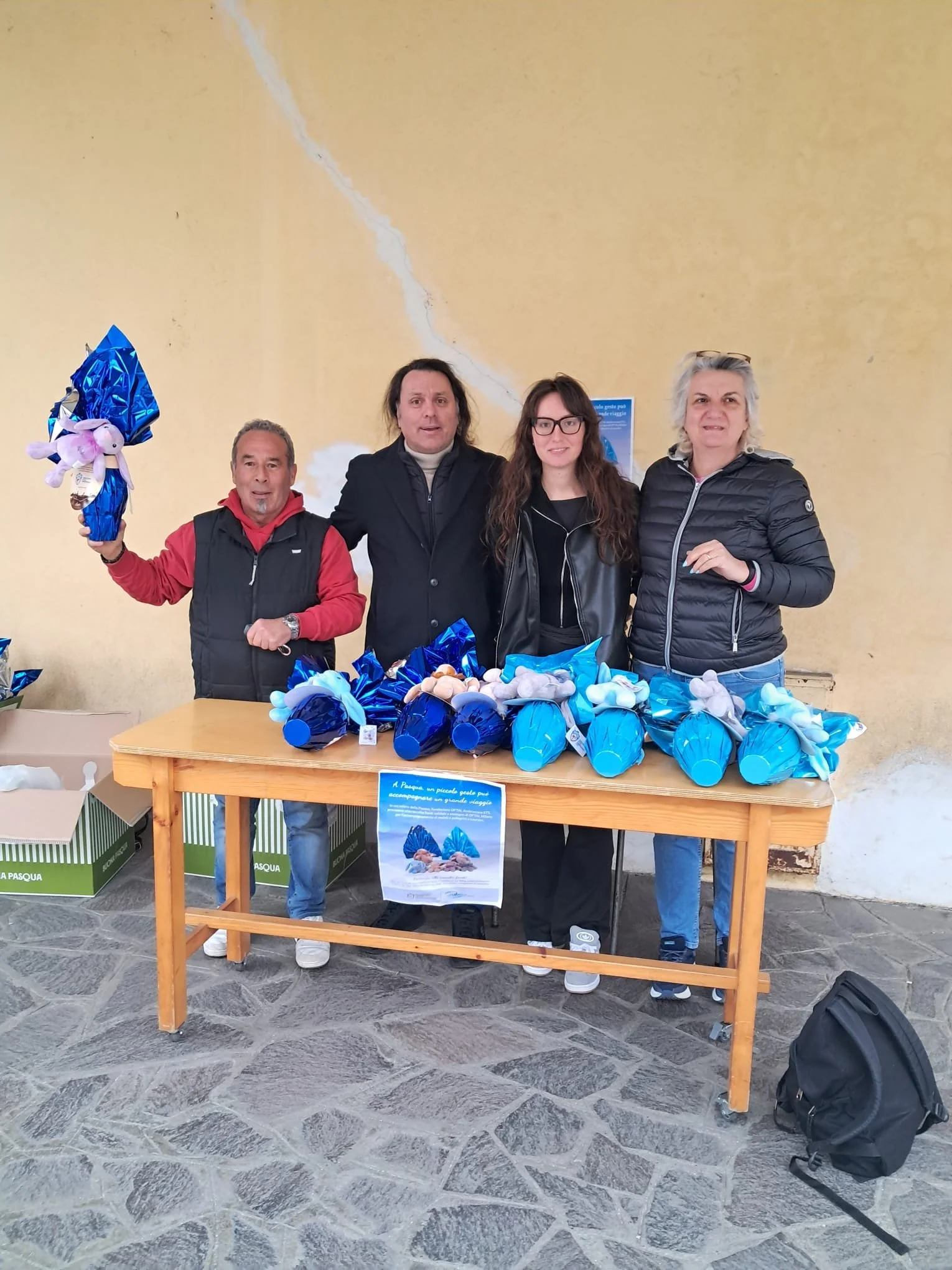 Gruppo di quattro persone che partecipano a una manifestazione pasquale. Sul tavolo ci sono uova di Pasqua decorate con materiali azzurri e in cartoncino, e qualche gadget. Dietro di loro un muro color crema con crepe visibili.