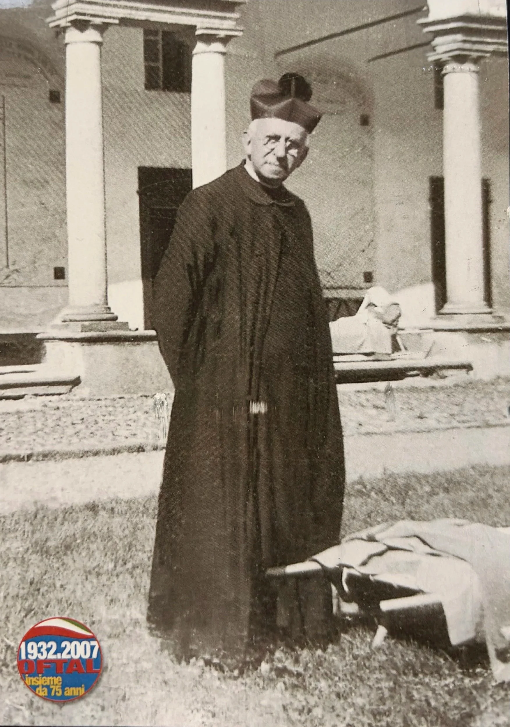 Foto in bianco e nero di un uomo vestito con un cappotto lungo e cappello gigante, davanti a un edificio con colonne. Sul lato inferiore sinistro, un logo celebrativo di 75 anni di attività con le date 1932-2007.