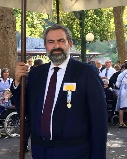 Uomo con barba e capelli grigi, vestito con abito scuro e cravatta, con medaglia appuntata al petto, che tiene un ombrello in un evento all'aperto con molte persone intorno.