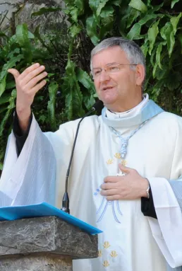 Prete che parla durante una cerimonia religiosa, indossa abiti liturgici e è circondato da piante verdi.