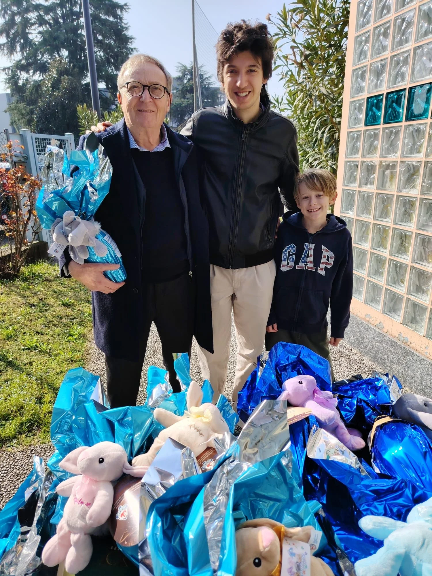 Tre persone, un anziano uomo e due giovani, sorridenti, in piedi davanti a un grande mucchio di bambole di pezza avvolte in sacchetti di plastica azzurri e argento, in un cortile esterno con siepi e una parete di vetro.