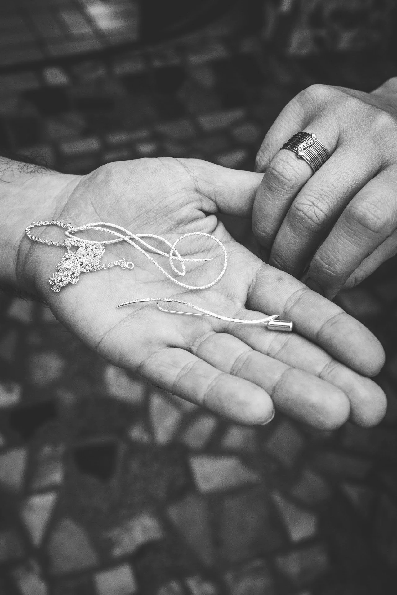 Mãos segurando joias de ouro, incluindo uma corrente, um colar com pingente, um brinco e uma aliança, em uma imagem em preto e branco com fundo de pedras.