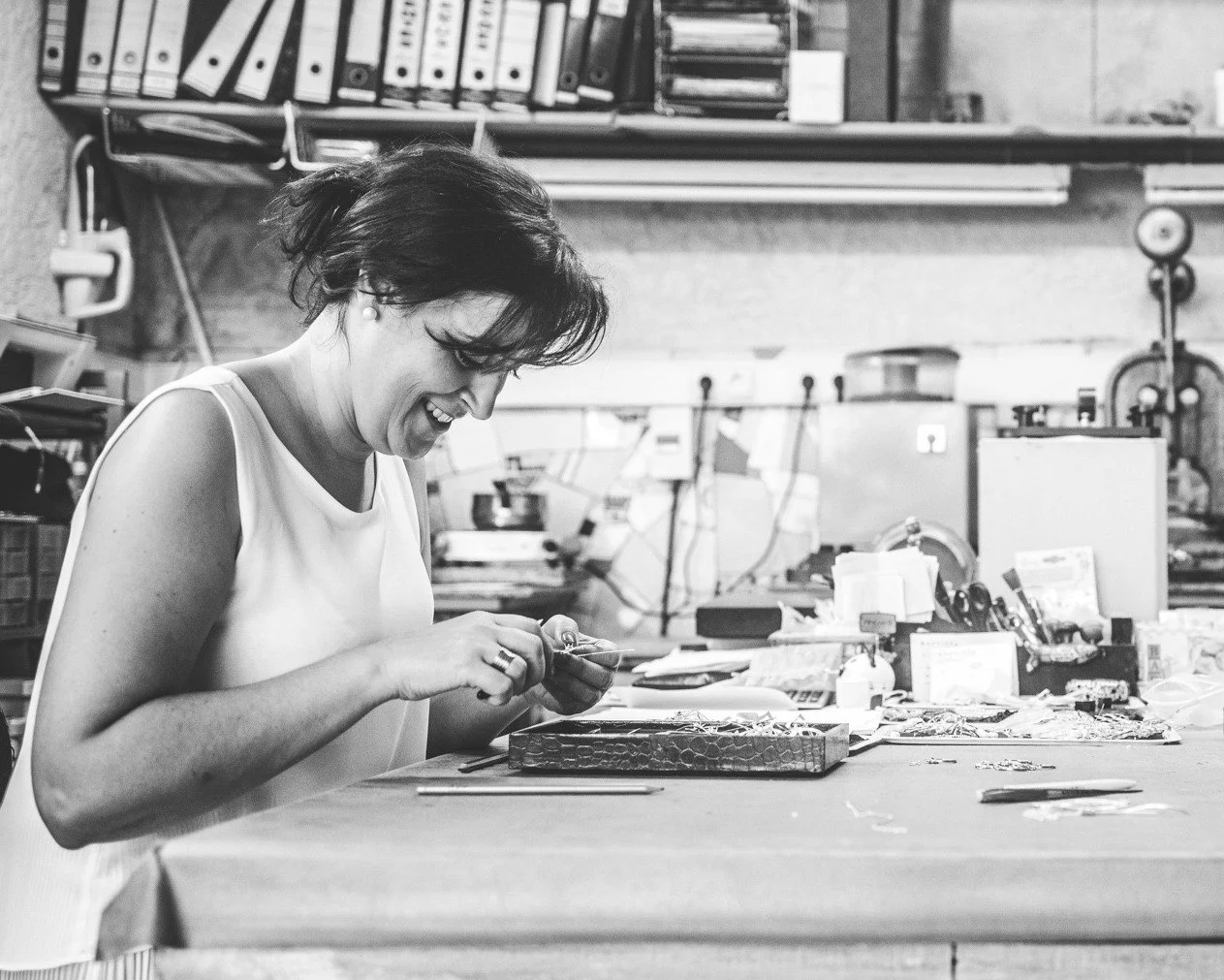 Mulher sorridente trabalhando em um ateliê de joalheria, manuseando uma peça de joalheria, com ferramentas e materiais espalhados na mesa