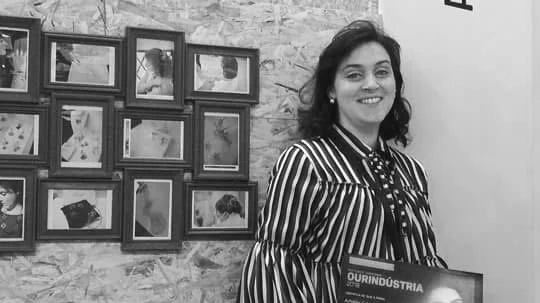 Mulher sorridente segurando uma placa, ao lado de uma parede com várias molduras com fotos.