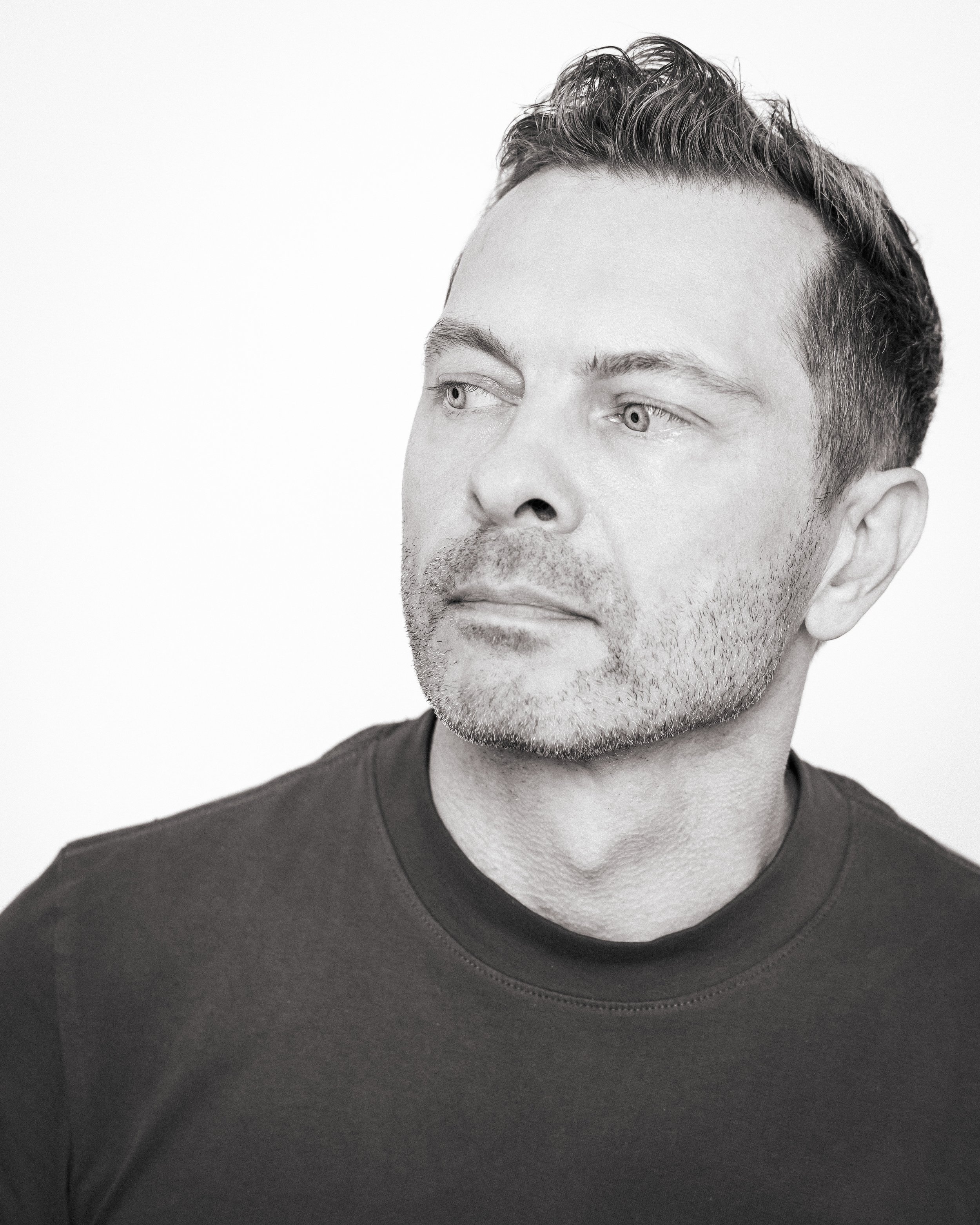 Black-and-white portrait of a man with short hair and stubble, looking to the side, wearing a dark T-shirt.