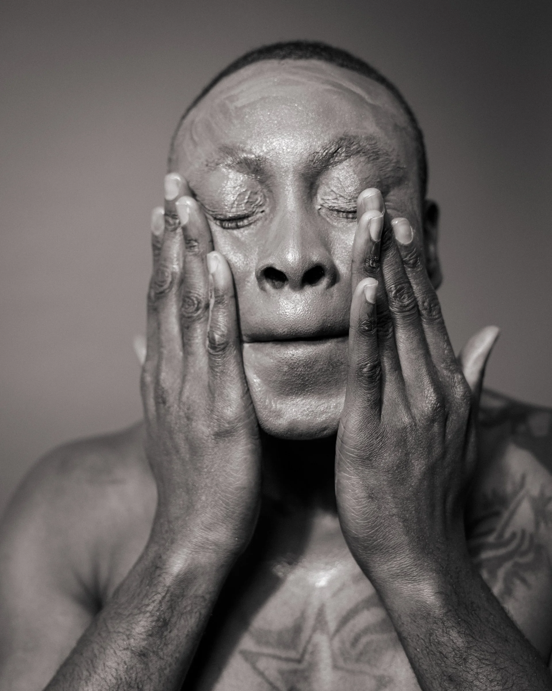 Black and white photo of a person with eyes closed, hands on face, skin texture visible, tattoo on chest.