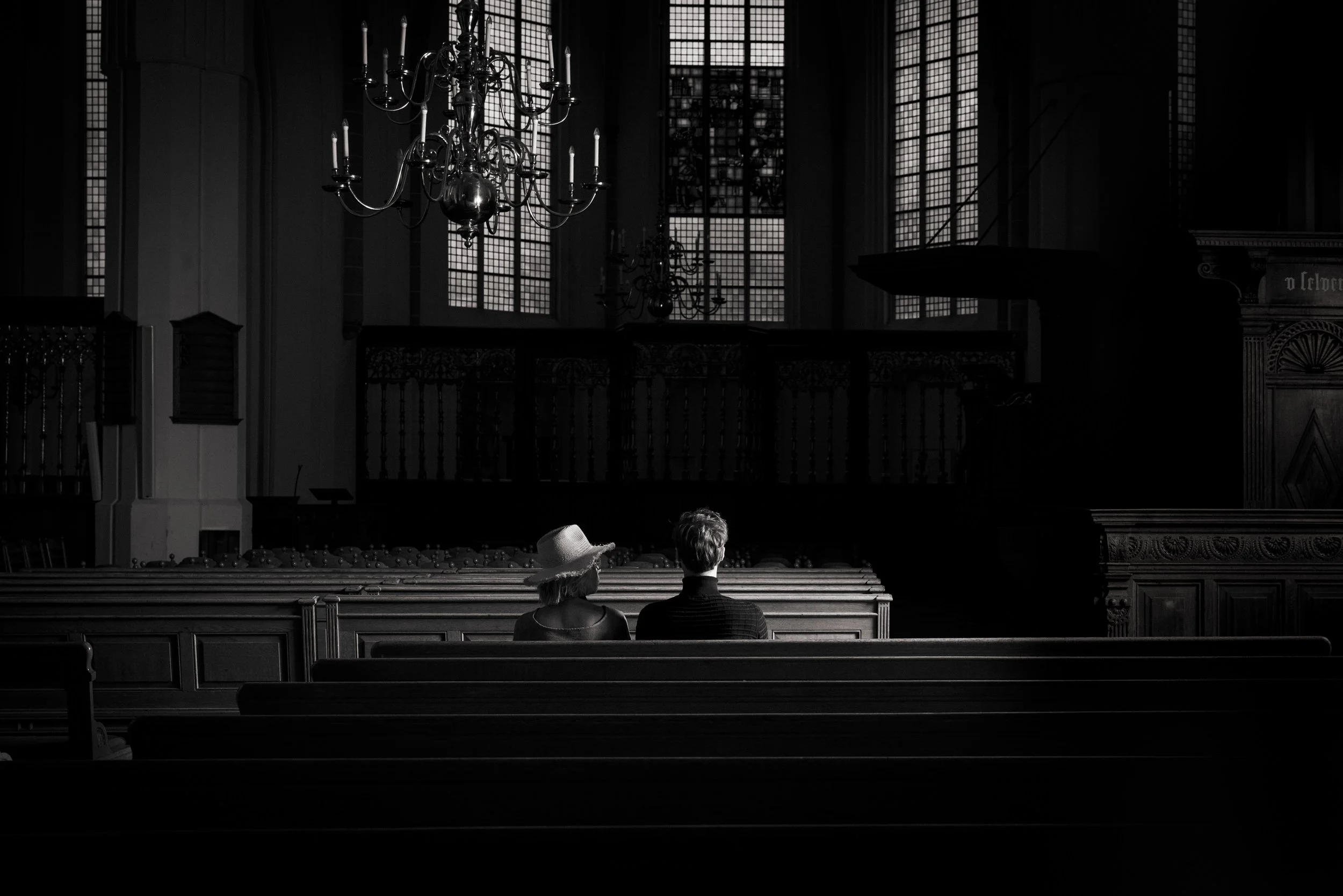 Two people sit on pews inside a church or cathedral with large stained glass windows. The scene is in black and white, with a chandelier hanging from the ceiling and intricate woodwork visible.