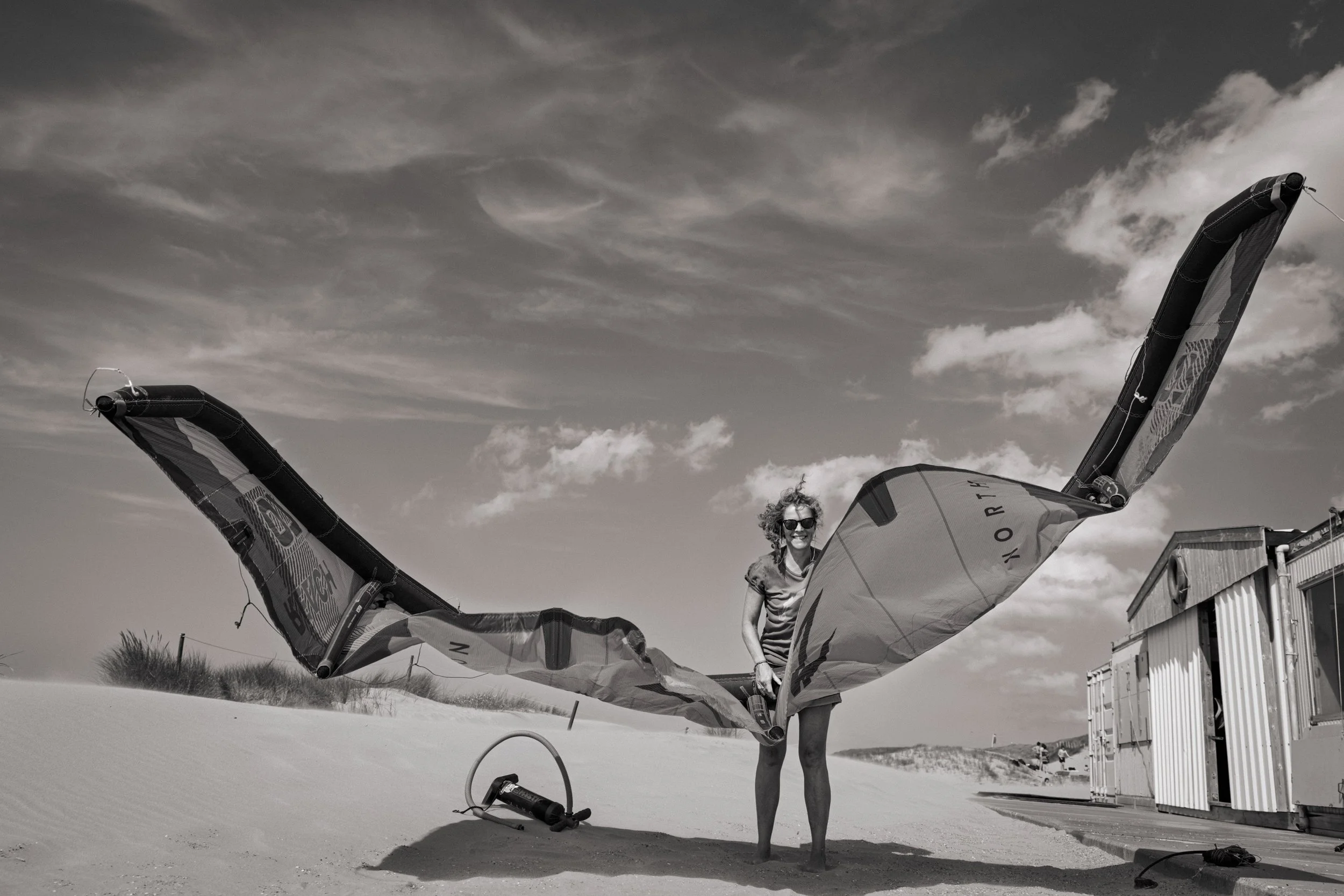 A woman standing on a sandy beach holding the wings of a kite. She is wearing sunglasses and a striped t-shirt, with a house or temporary building in the background under a sky with scattered clouds.