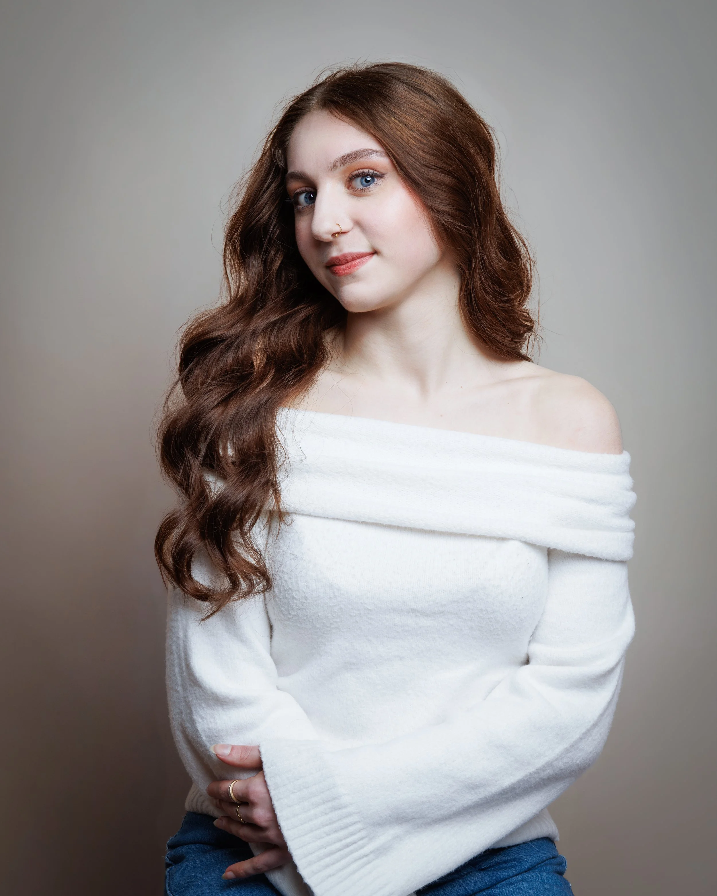 Young woman with long, wavy reddish-brown hair wearing a white off-shoulder sweater and sitting against a plain background.