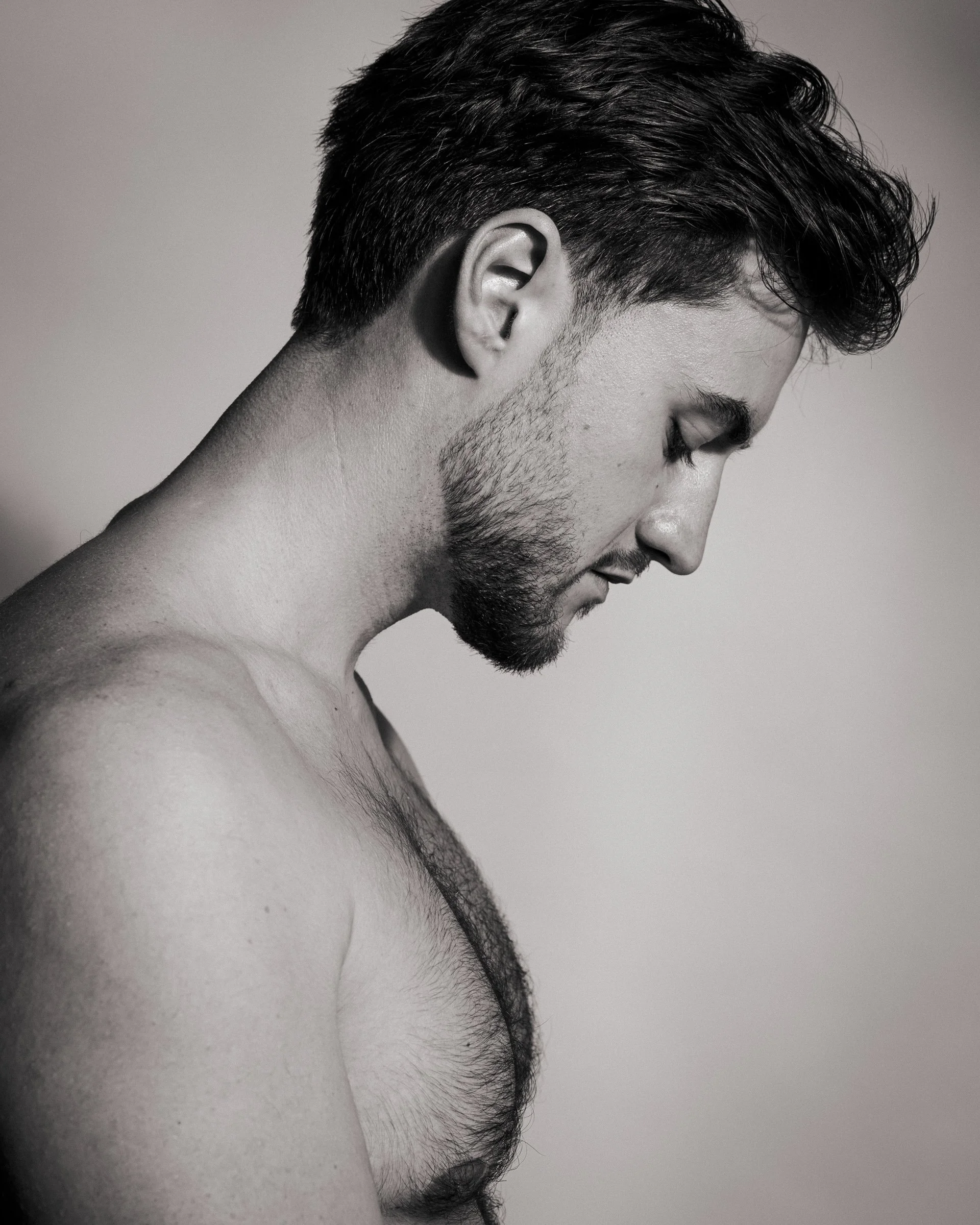 A black-and-white side profile of a shirtless young man with dark hair and a beard, looking downward.